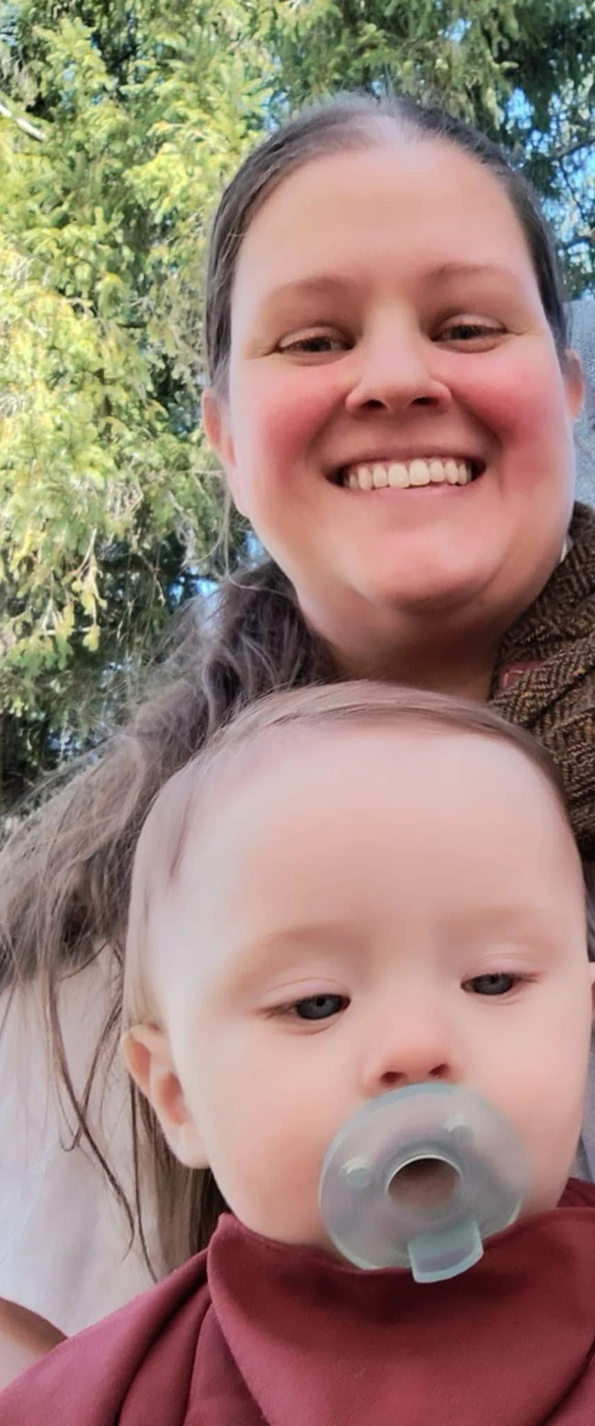 A woman smiling outdoors with a young child, who has a pacifier, in front of her, with trees and blue sky in the background.