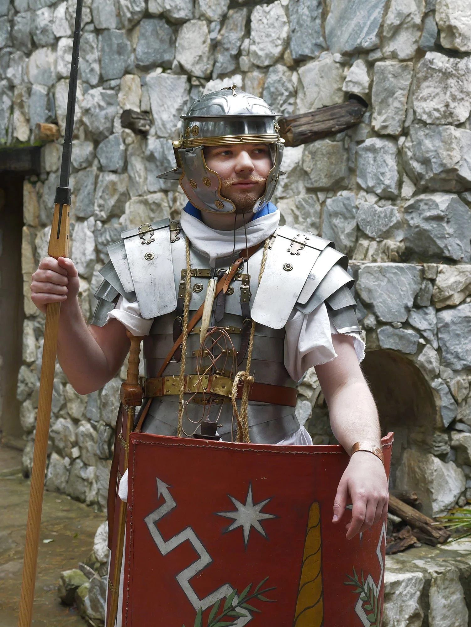 A person dressed as a Roman soldier in armor with a helmet and a shield, holding a spear, standing in front of a stone wall.
