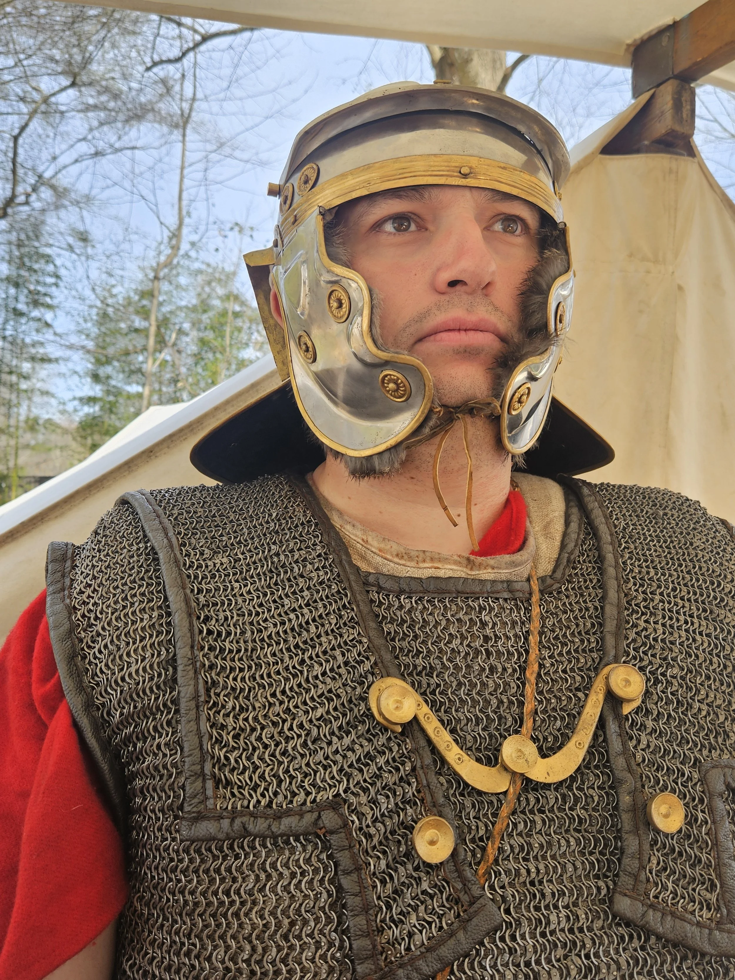 Person dressed as a Roman soldier wearing a metal helmet with gold accents, chainmail armor, and a red shirt underneath. Outdoors with trees and a tent in the background.