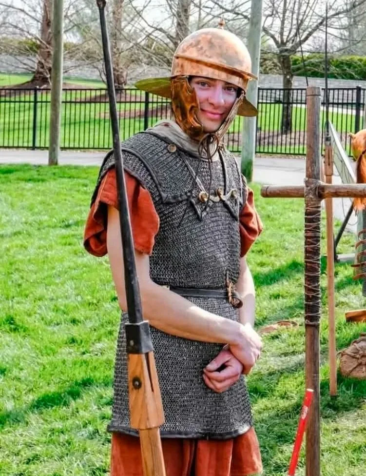 Person dressed as a medieval soldier, wearing chainmail armor, a helmet, and holding a spear, standing outdoors on a grassy area with a fence and trees in the background.