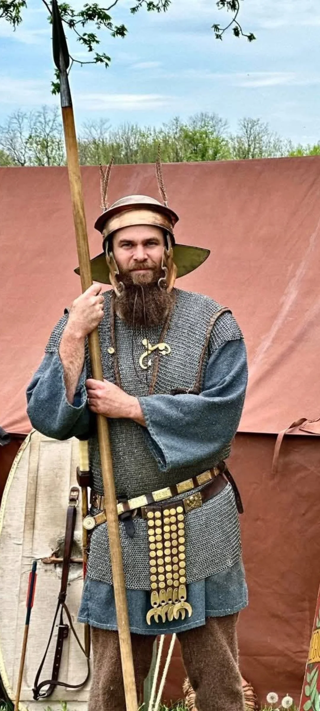A man dressed as a medieval warrior or Viking, holding a spear, wearing chainmail armor, a helmet with animal horns, and a large belt with decorative hooks, standing outdoors in front of a brown tent.