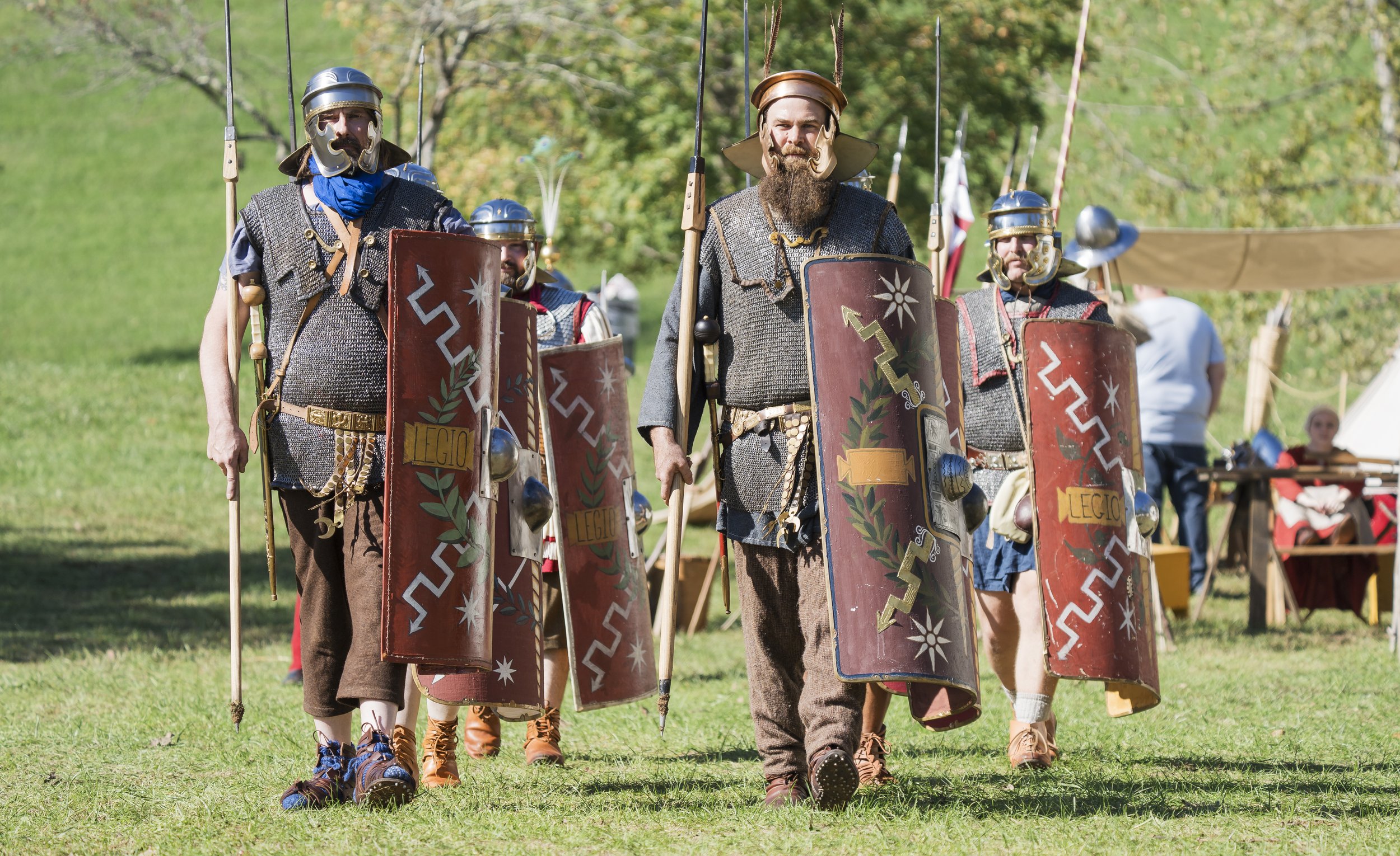 Group of men dressed as Roman soldiers participating in a historical reenactment outdoors, holding shields and spears, with a grassy field and trees in the background.