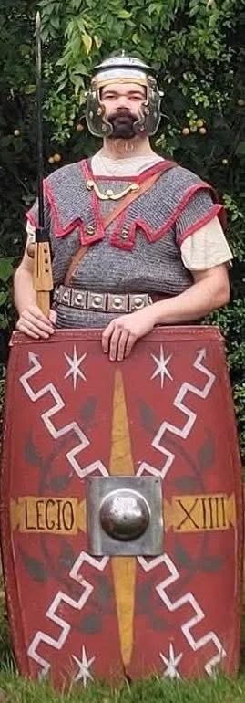Person dressed as a Roman soldier holding a shield with Roman numerals and symbols, standing outdoors in front of greenery.