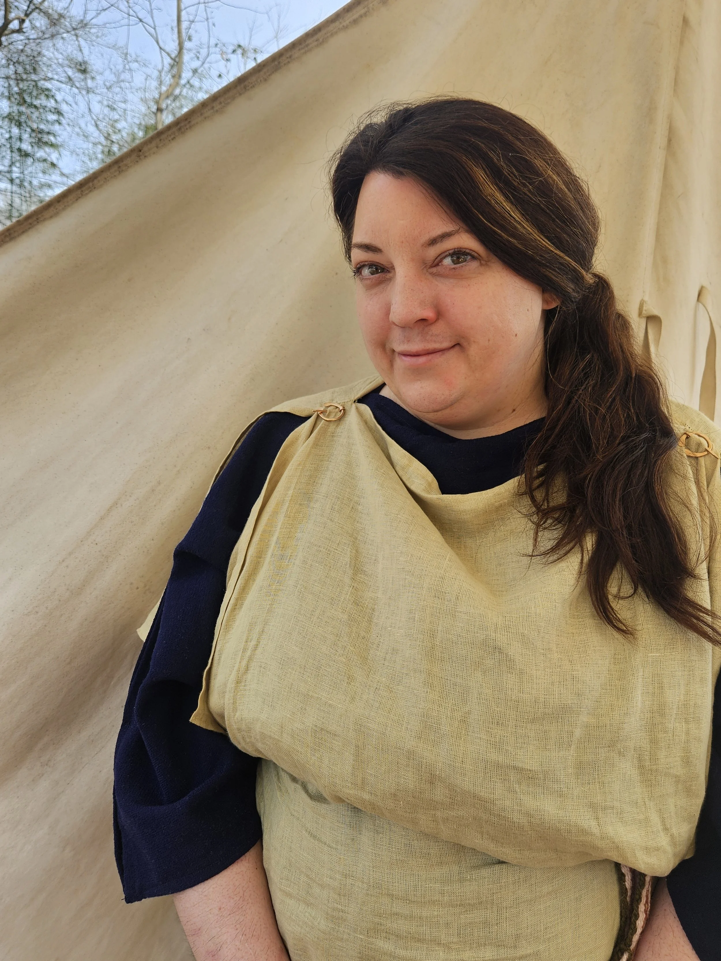 A woman with brown hair and blue eyes smiling at the camera, wearing a navy blue top underneath a beige garment, standing outdoors against a light-colored fabric or canvas backdrop, with some trees and blue sky visible in the background.