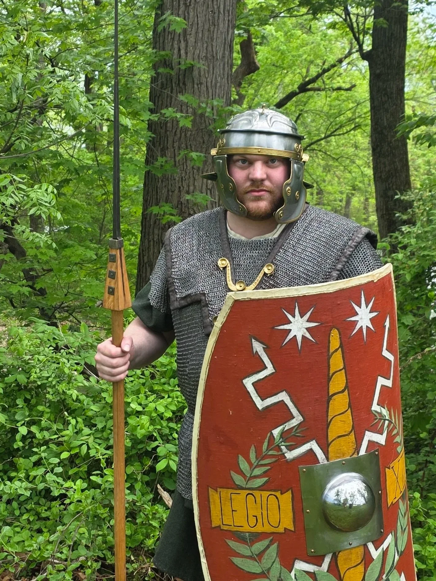 A man dressed as a Roman soldier holding a spear and shield in a forest with green foliage.
