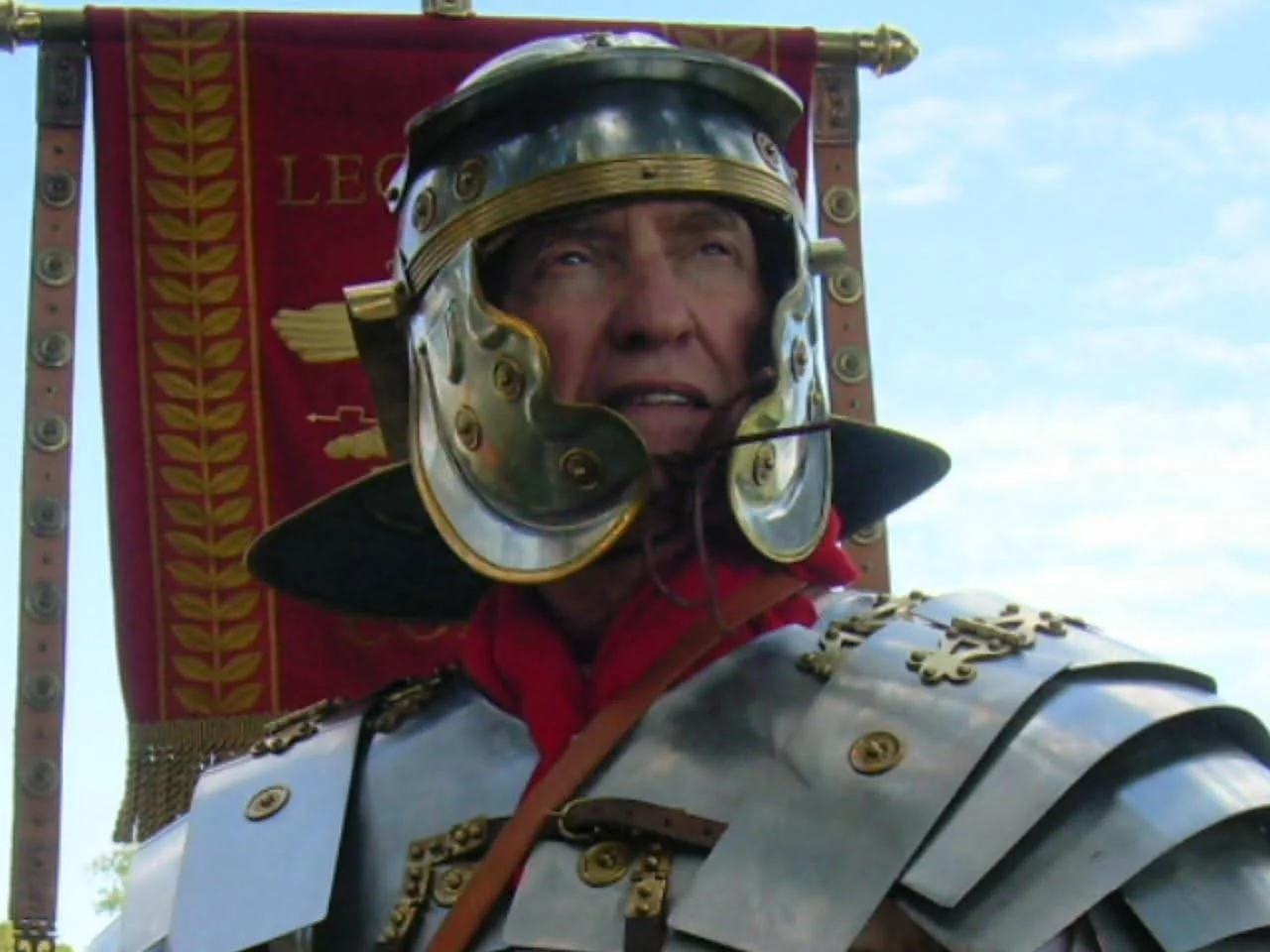 A man dressed as a Roman soldier, wearing a silver helmet with gold details, armor, and a red cape, standing in front of a red banner with gold laurel leaves and the word 'LEO'.