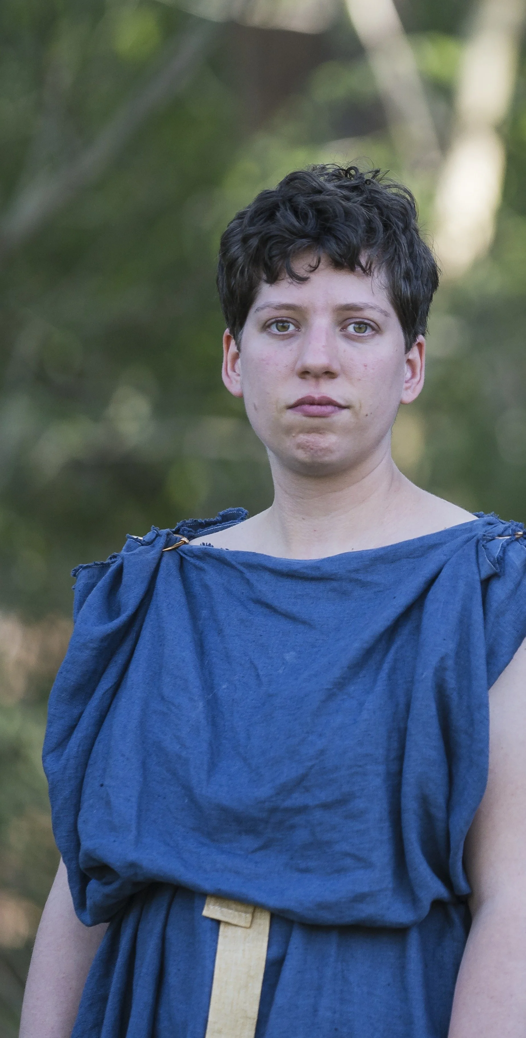 A woman with short curly dark hair and fair skin standing outdoors with a blurred green background, wearing a loose blue garment.