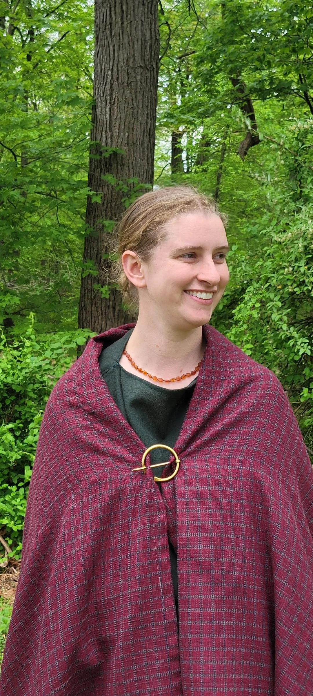 A woman with light brown hair smiling in a green forest, wearing a dark shirt and a red plaid cloak fastened with a gold brooch.