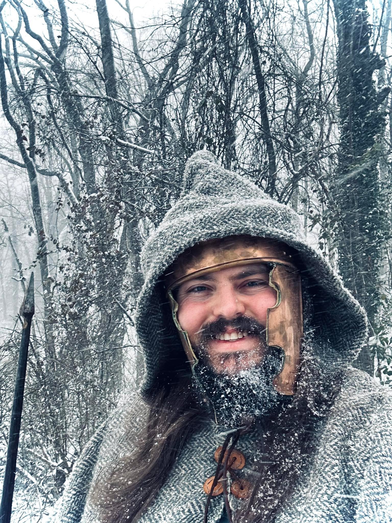 A man with a beard and long hair, smiling, is dressed in winter clothing including a hooded coat and a wool hat in a snowy forest. Snow covers his face and clothing, with a spear visible in the background.