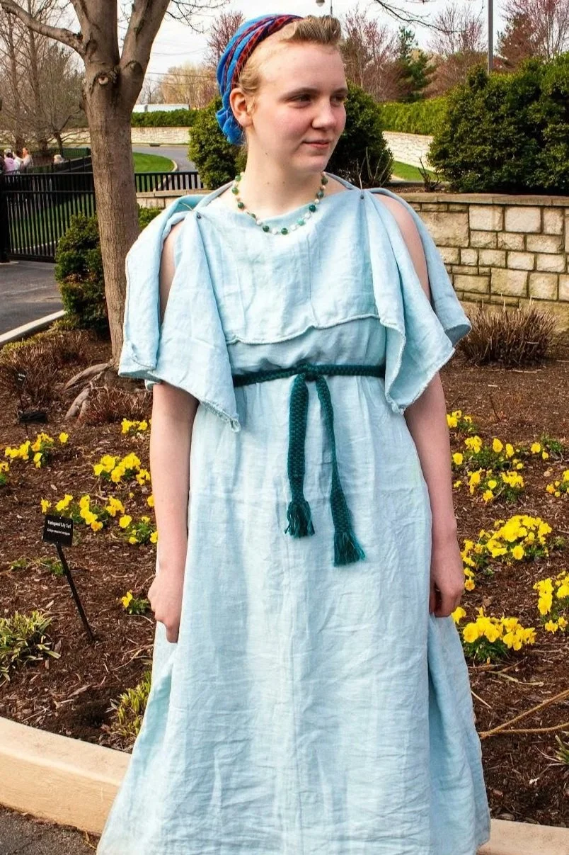 A young woman standing outdoors near a flower garden, wearing a light blue historical dress with shoulder cutouts, a dark green waist belt, a multicolored headscarf, and a necklace.
