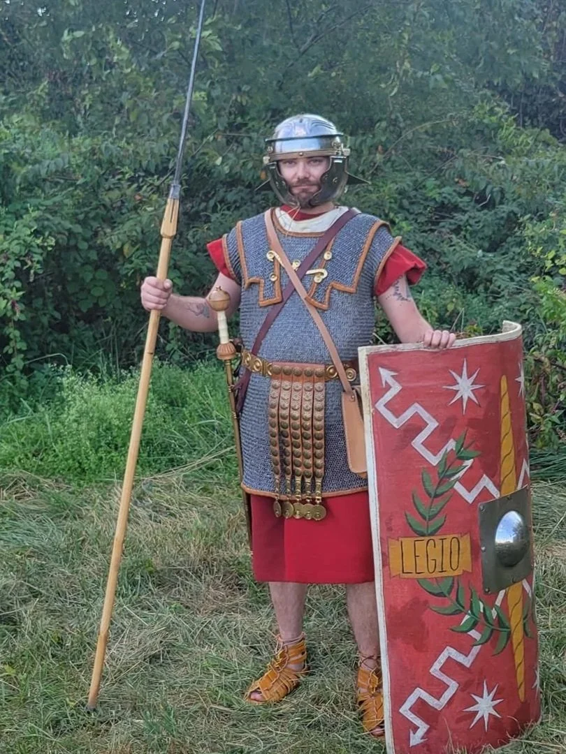 Person dressed as a Roman soldier holding a spear in one hand and a shield labeled 'LEGIO' in the other, standing outdoors on grass with greenery in the background.