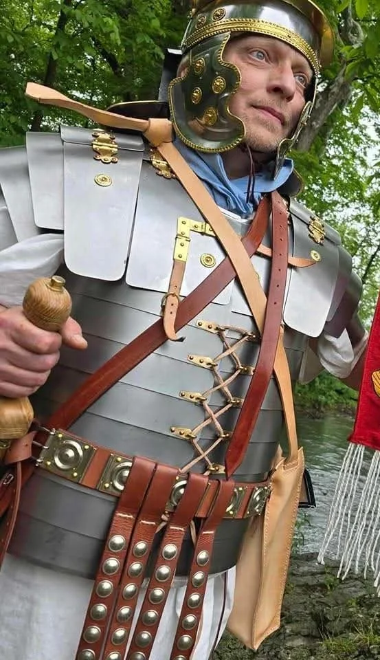 A man dressed as a Roman centurion in metal armor and a helmet, holding a wooden sword, standing outdoors with green trees in the background.