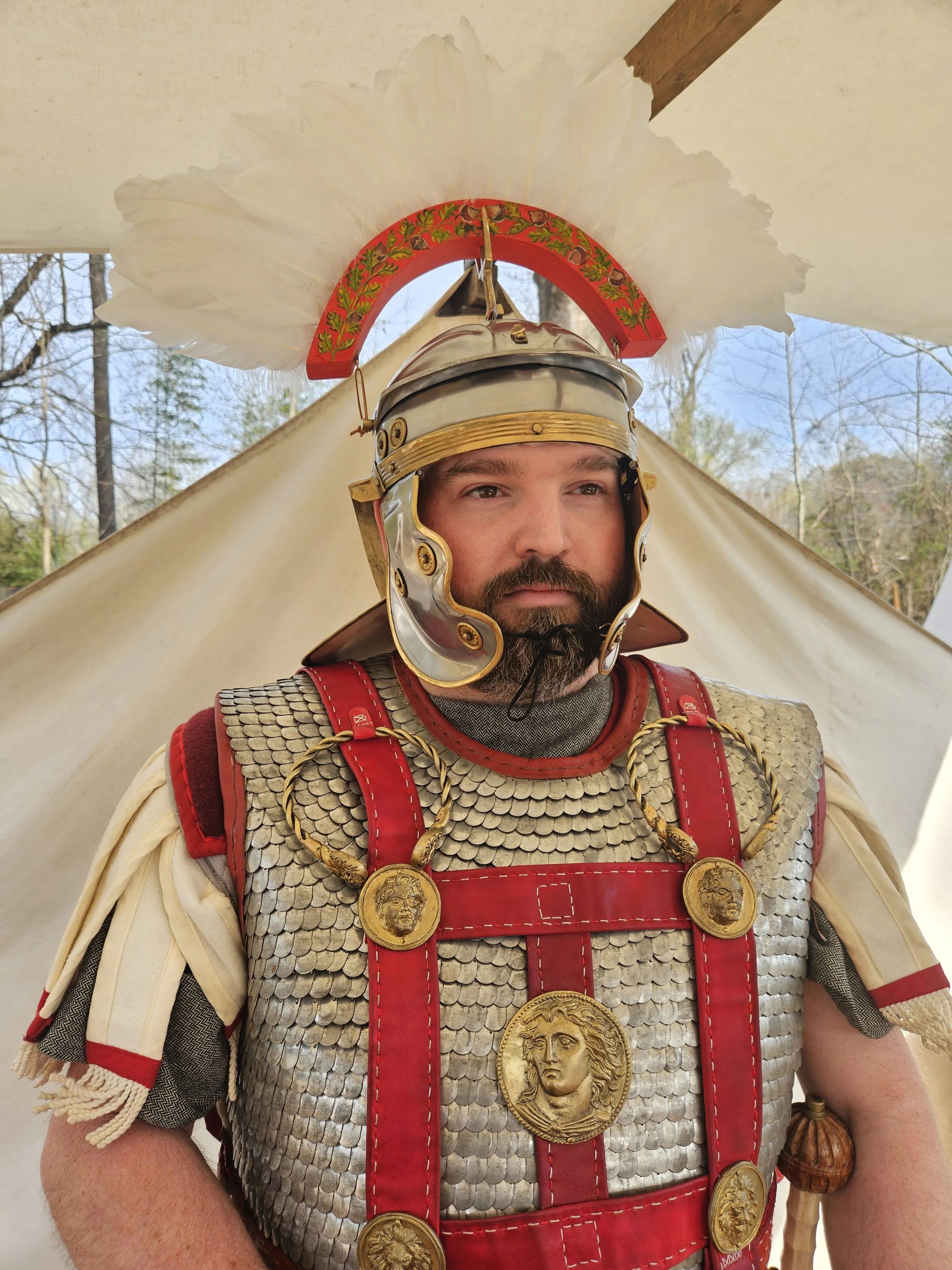A man dressed as a Roman soldier, wearing a helmet with a feathered crest and chainmail armor with red accents, standing outdoors near a tent with trees in the background.