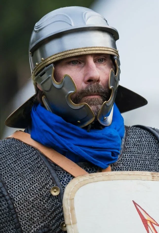 A man dressed as a medieval knight with a helmet, chainmail armor, a blue scarf, and holding a shield.