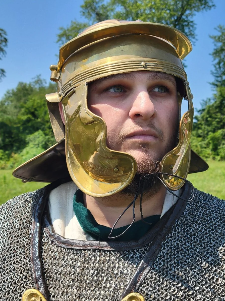 Close-up of a man wearing a medieval-style chainmail shirt and a brass helmet, outdoors with green trees and blue sky in the background.