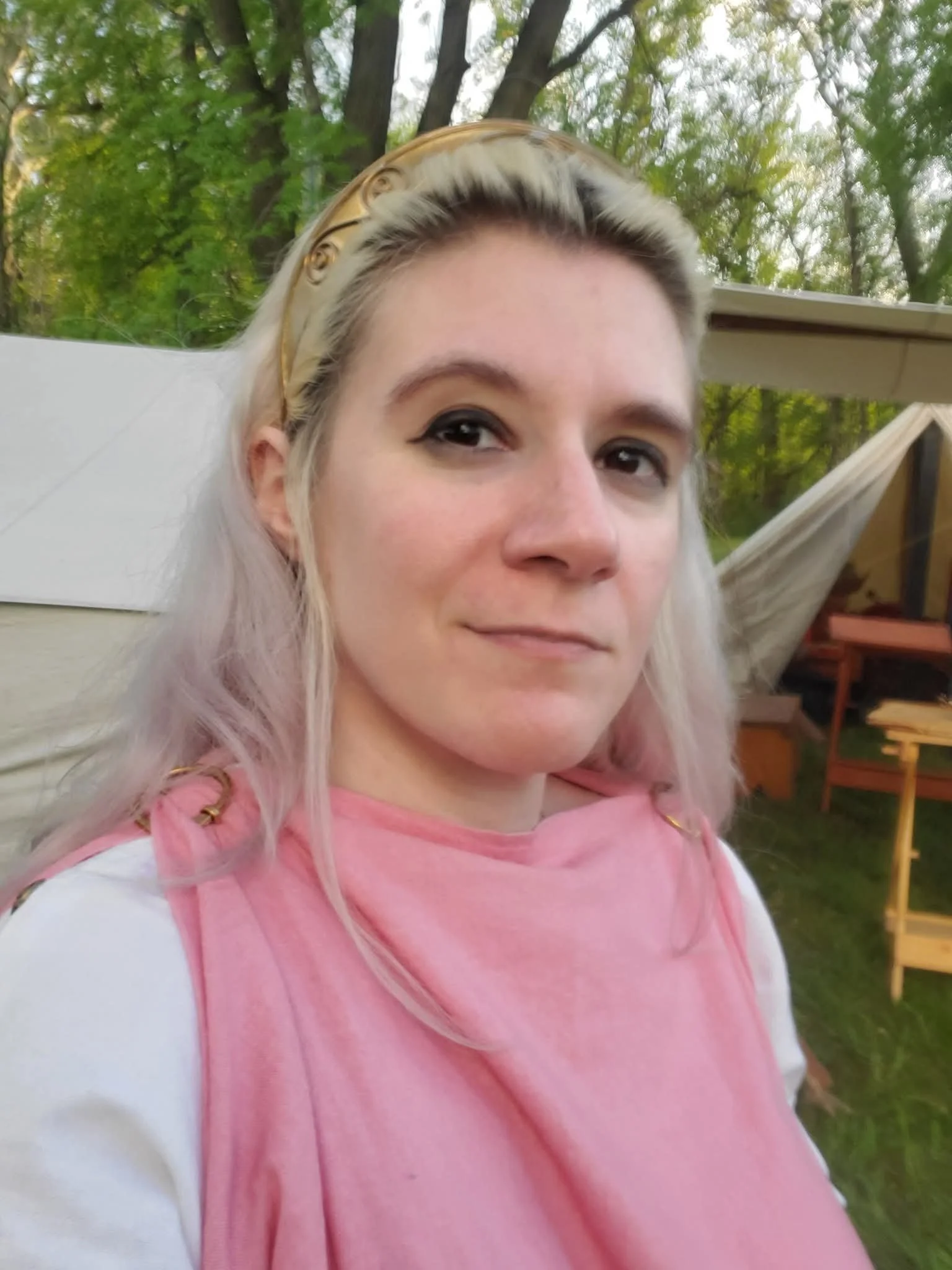 A woman with platinum blonde hair and a gold headband takes a selfie outdoors, with trees and a backyard setup in the background.