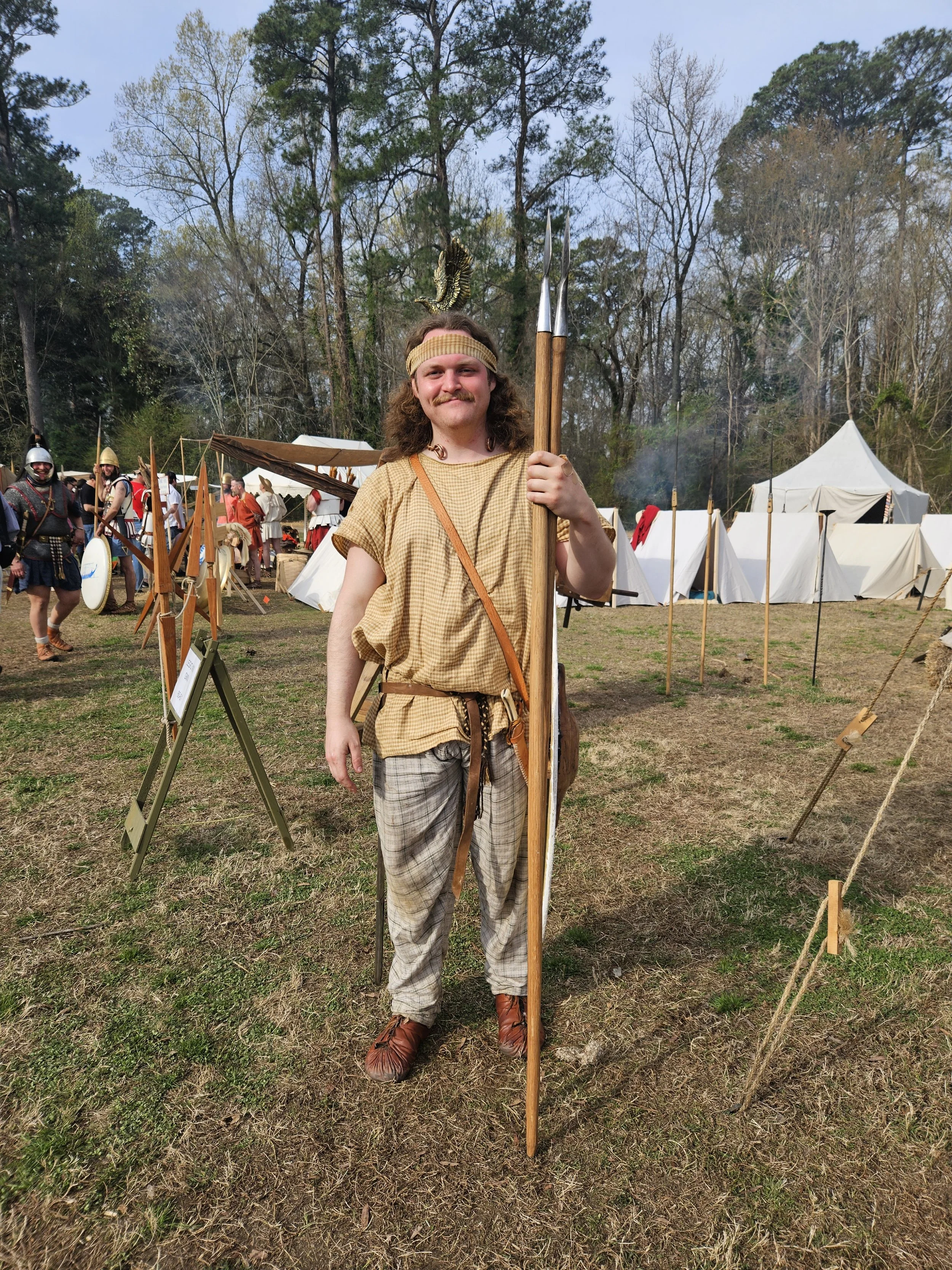 Man dressed in medieval costume, holding a spear, at a historical reenactment event with tents and other participants in medieval attire in the background.
