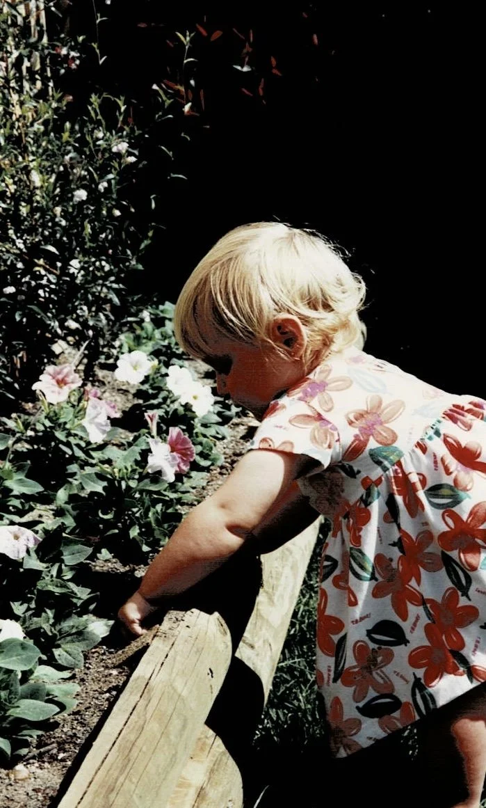 A blonde-haired girl in a floral dress leaning over a garden bed of pink and white flowers, with her arm resting on a wooden beam.