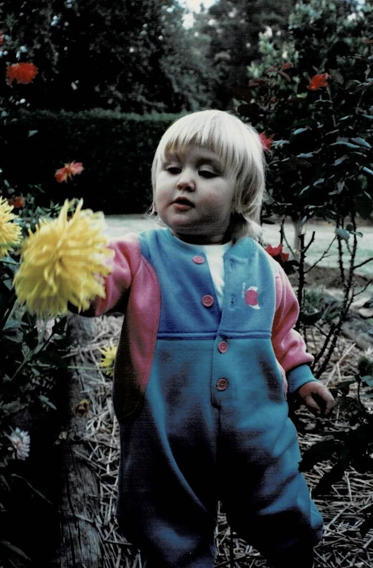 Young child with blonde hair wearing a colorful jacket, reaching out to touch a yellow flower in a garden surrounded by bushes and other flowers.