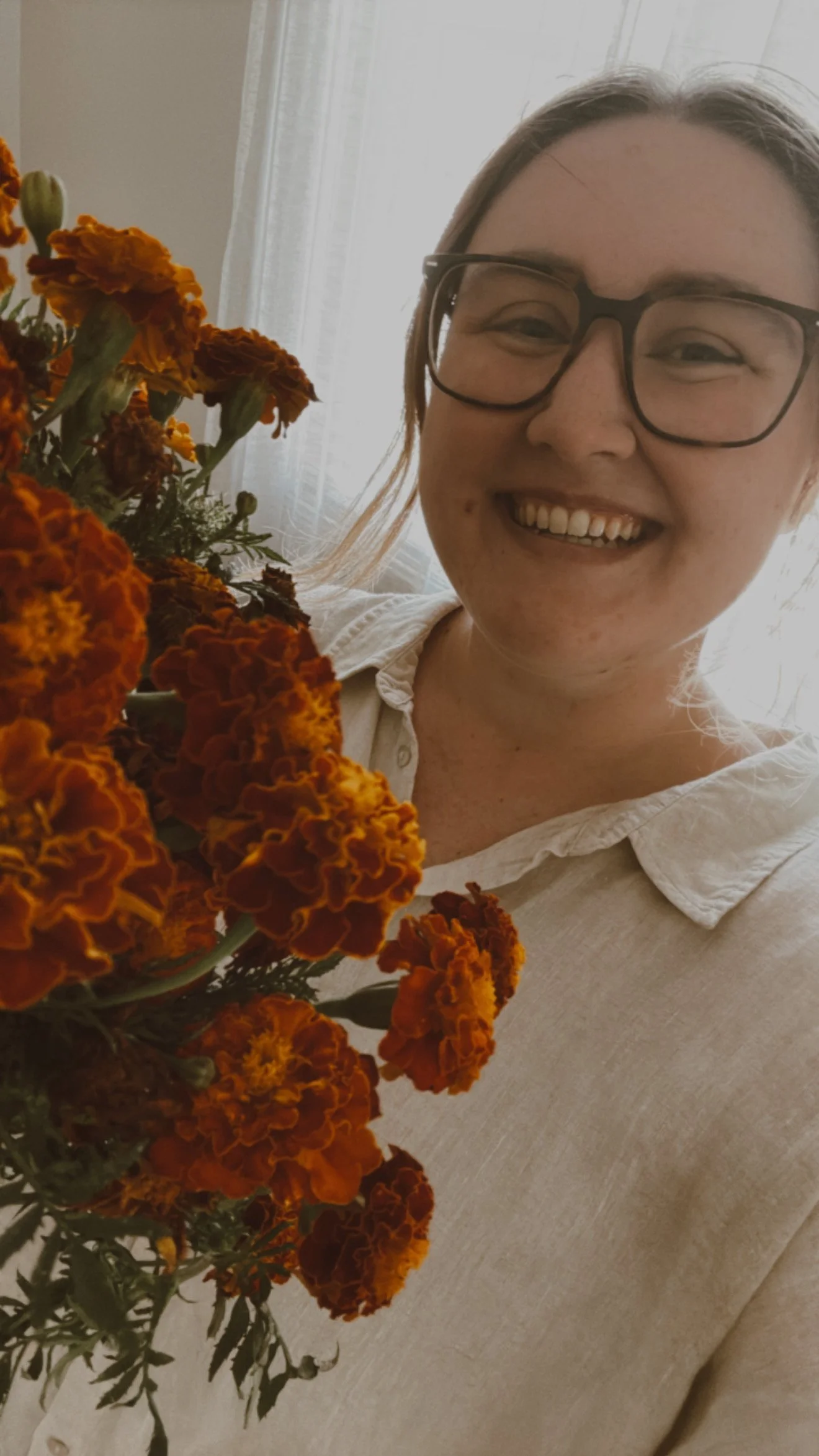 A woman smiling, holding a bouquet of orange marigold flowers indoors with sunlight coming through a window.