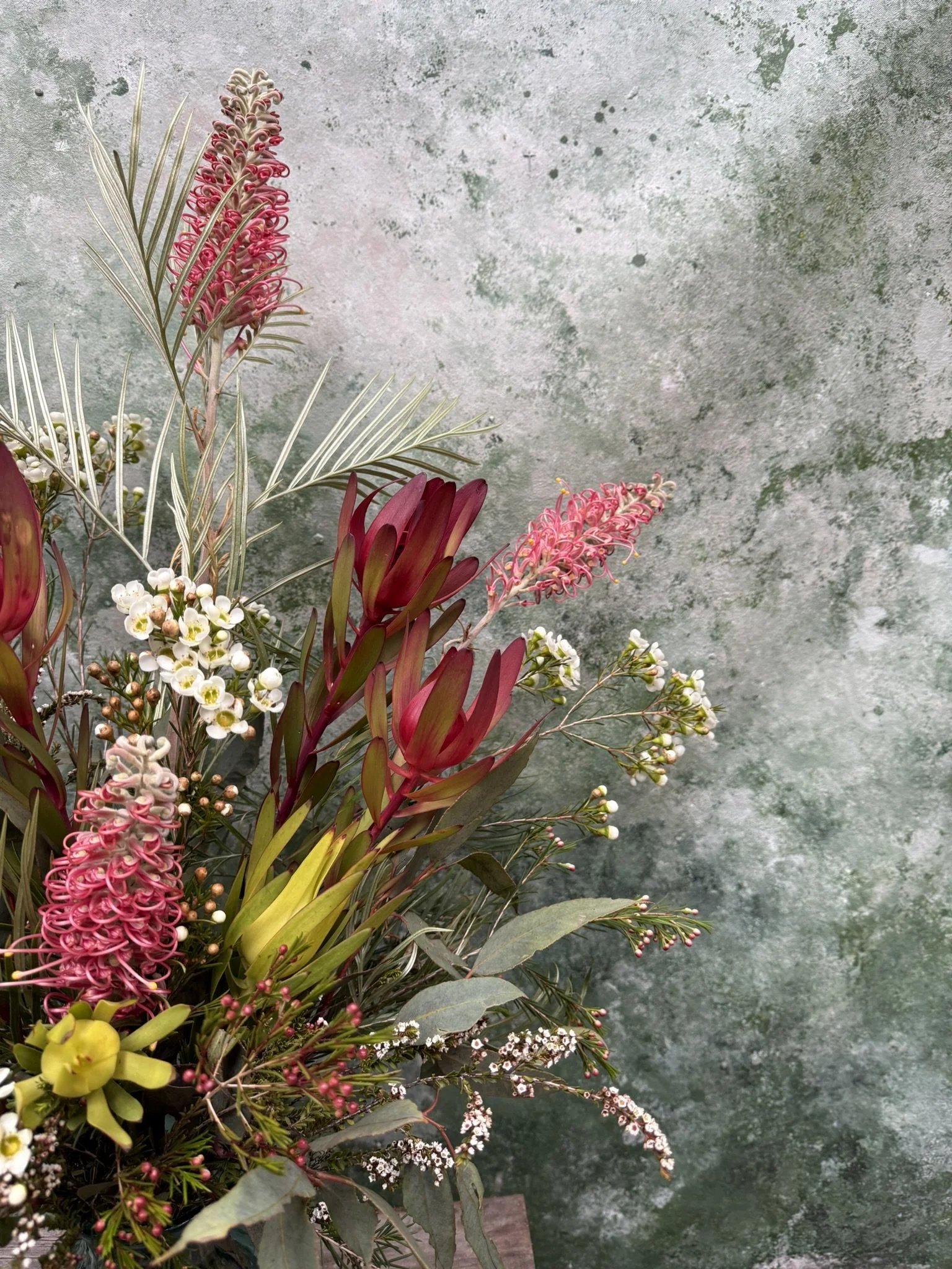A bouquet of pink, white, yellow, and green flowers and foliage against a textured green wall. Australian native wildflowers.