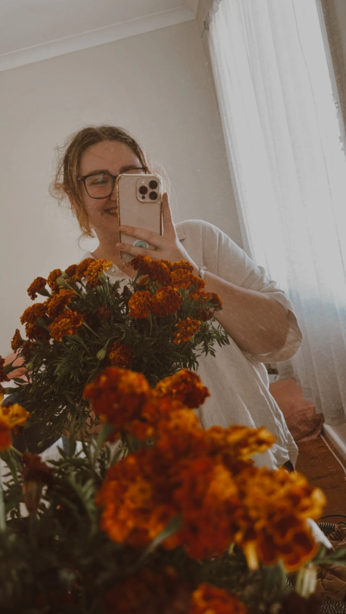 A person with glasses taking a mirror selfie in a room with flowers in the foreground and sunlight coming through a white curtain.