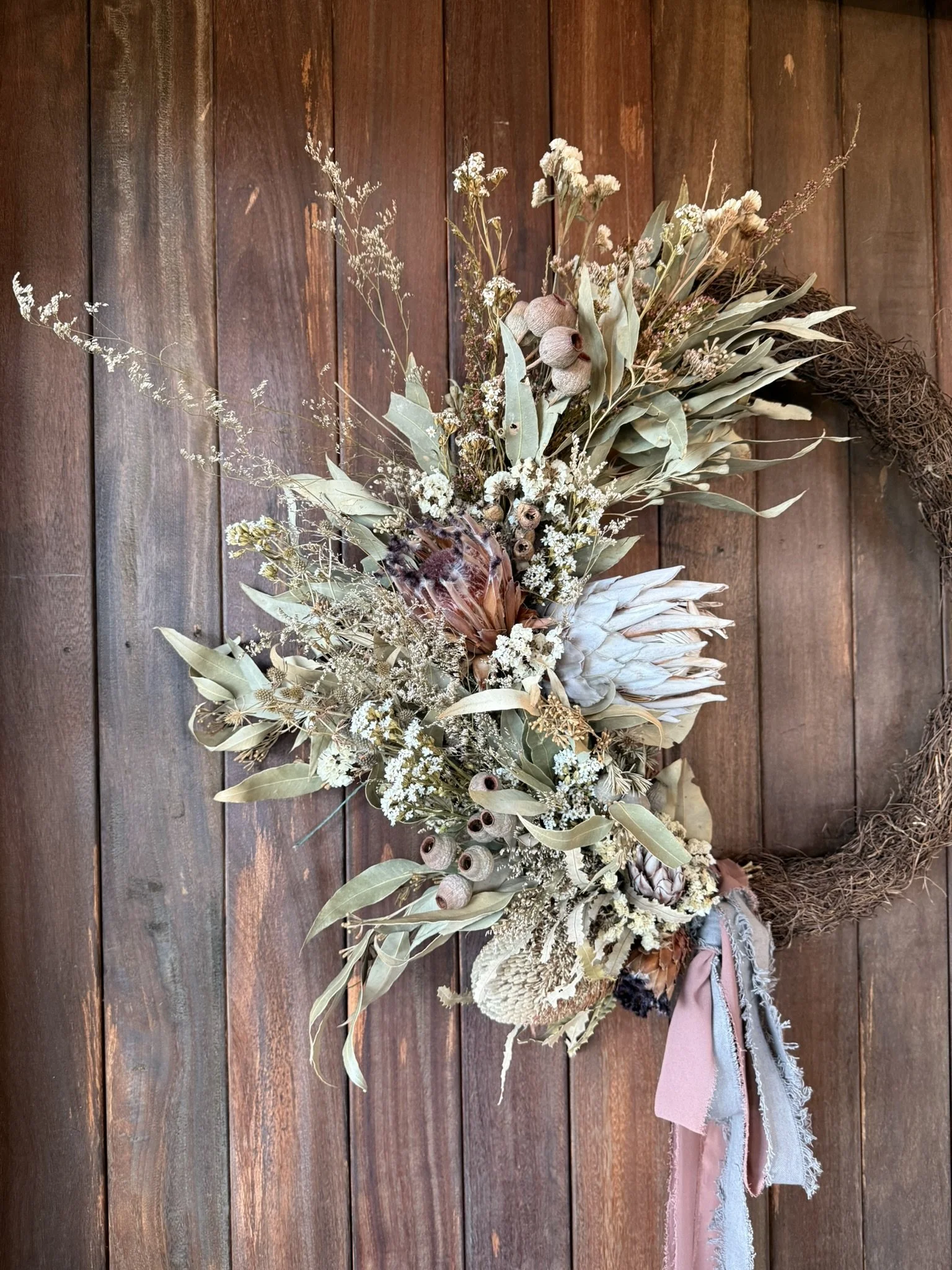 Dried floral wreath hanging on wooden wall with pink and beige fabric ribbons.