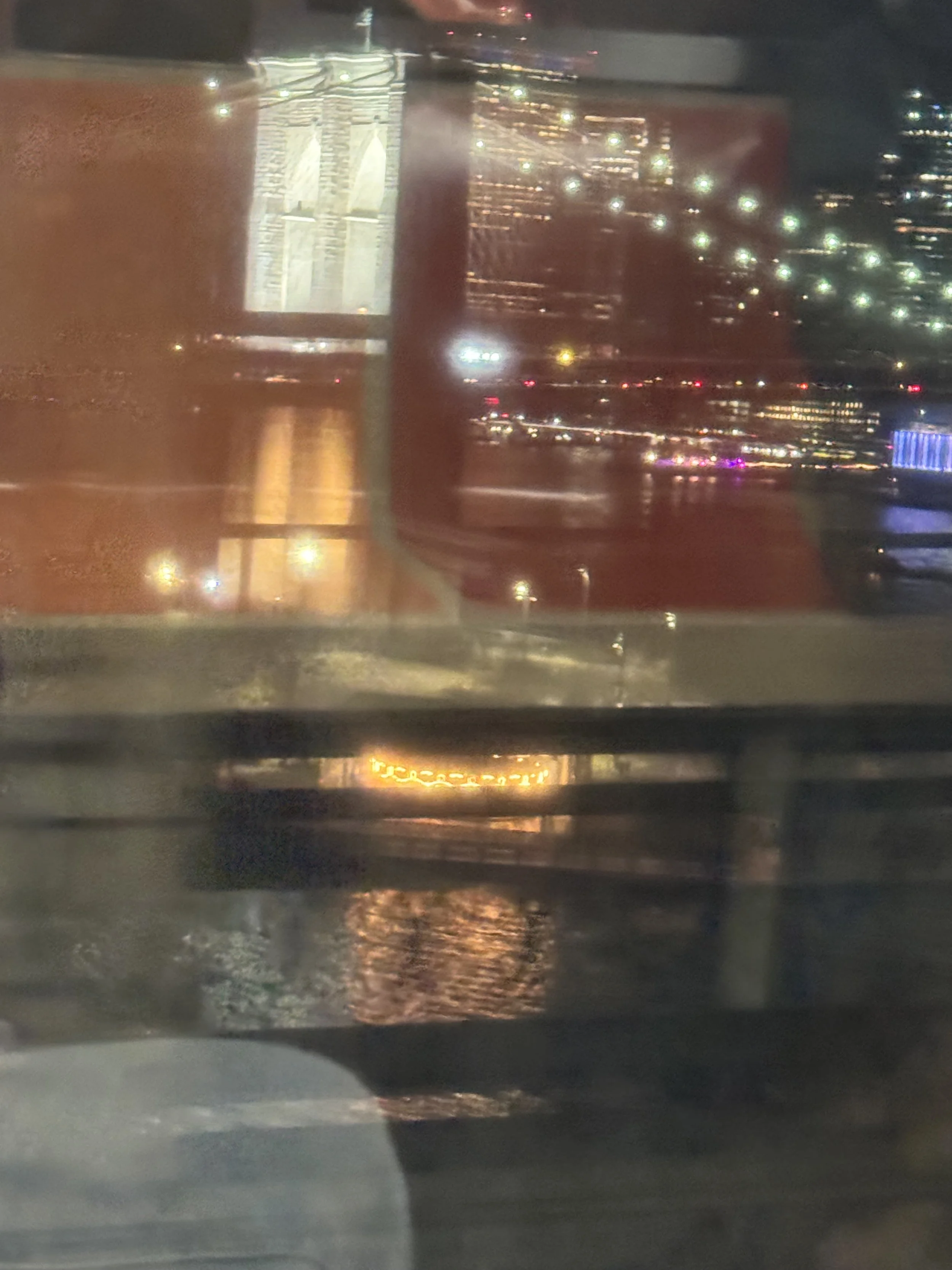 Nighttime cityscape seen through a window with reflections, featuring illuminated buildings, streetlights, and a river or body of water with reflections of city lights.