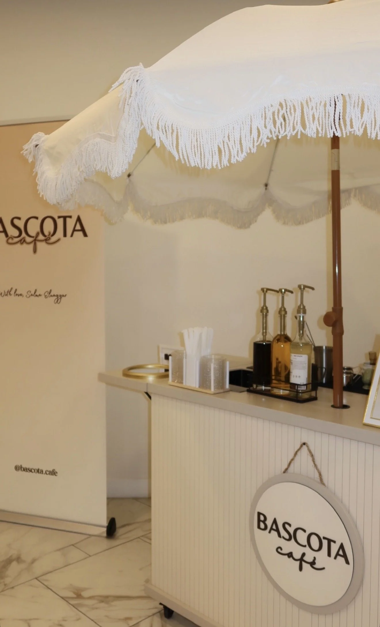 A white coffee bar with the logo 'Bascota Cafe' on the front, topped with a white table umbrella with fringe, situated in a cafe with marble tile flooring. Behind the bar, there are various bottles, a napkin dispenser, and a framed menu.