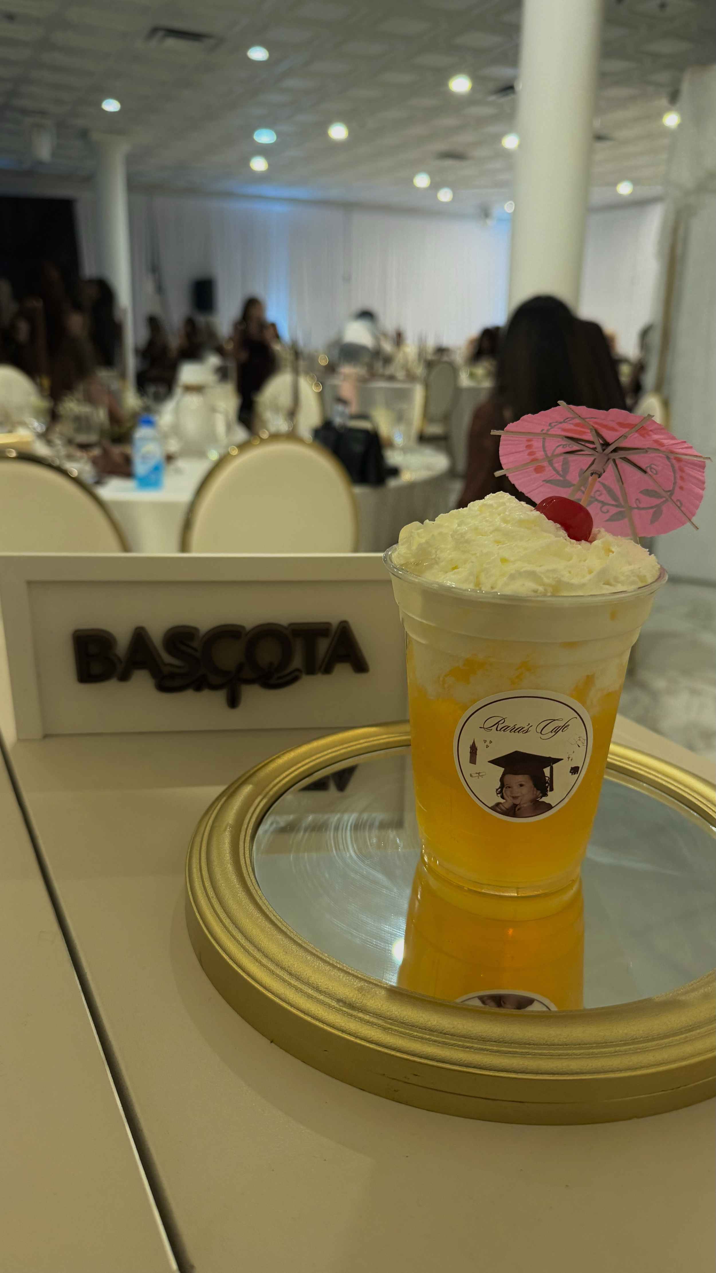 A tropical drink with whipped cream, a cherry, and a pink umbrella, on a gold tray in a banquet hall with people sitting at tables in the background.