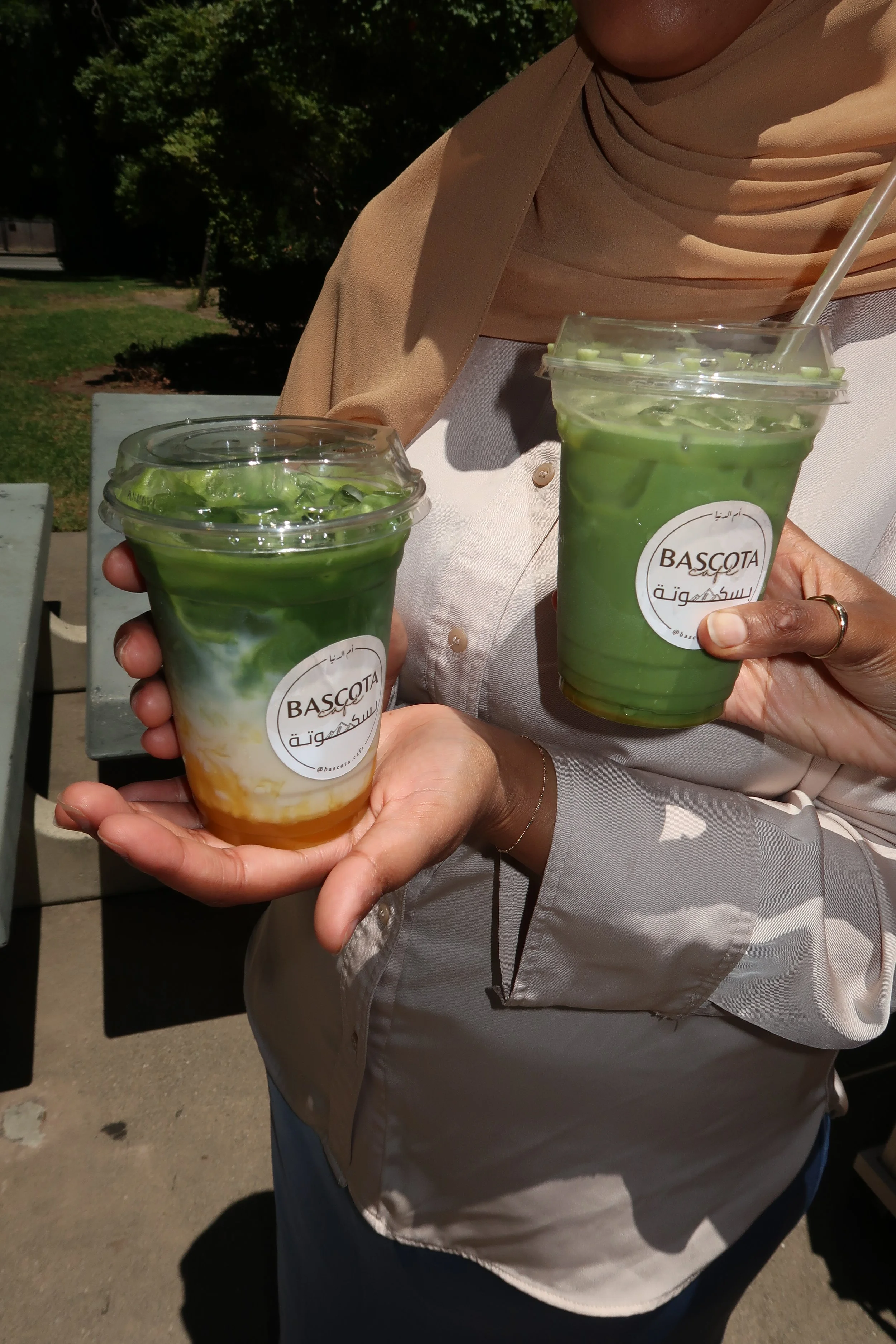 Two people holding cups of green matcha drink with white drizzle, labeled 'BASCOTA', outdoors on a sunny day.