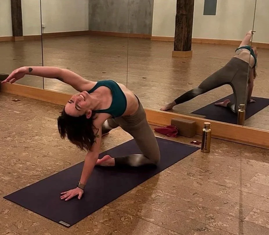 Corrie practicing yoga in a studio, performing a stretch on a black mat with a mirror reflecting her pose. She is smiling and her left arm is extended overhead, with her right knee on the mat.