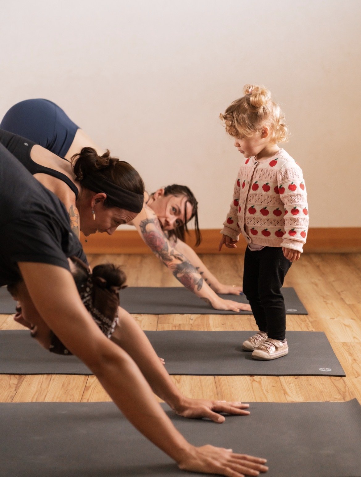 A group of students and a young girl practicing yoga on mats in a room with wooden floors. The students are in downward facing dog while the girl stands observing.