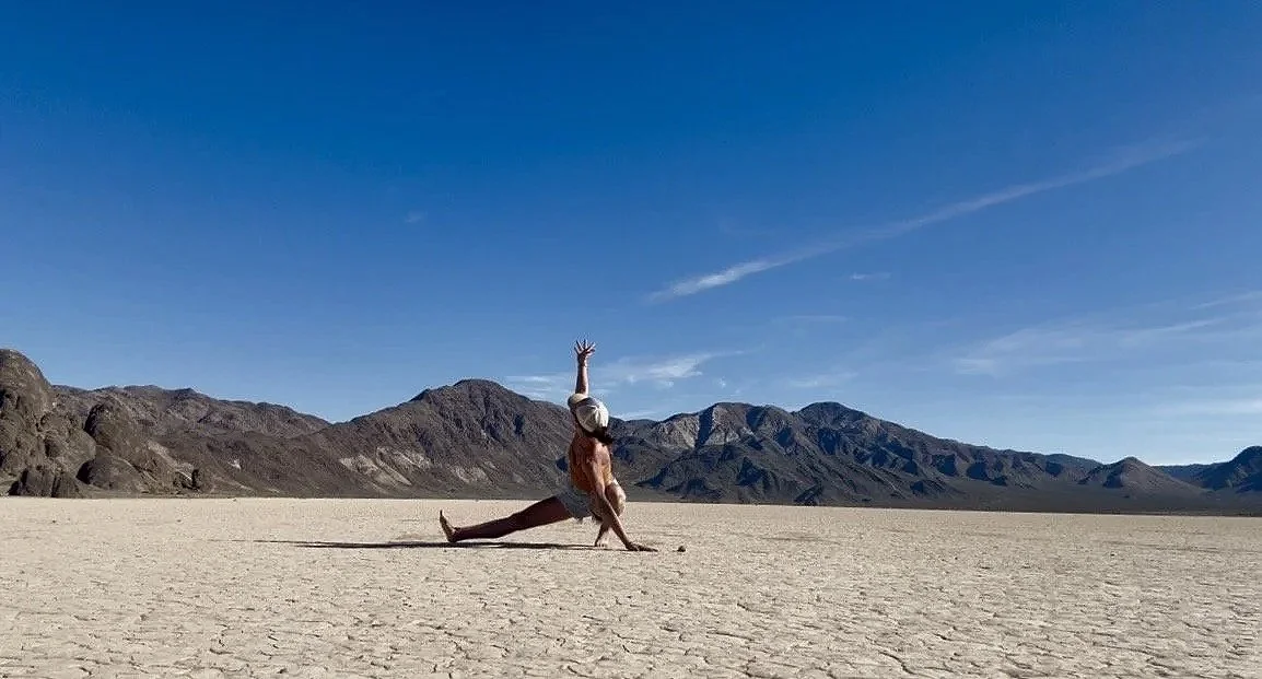 Corrie practicing yoga on a dry, cracked desert landscape with mountains in the background under a clear blue sky.