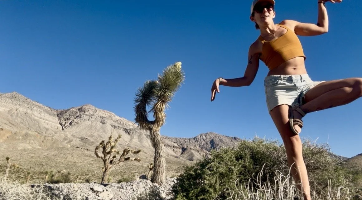 A woman levitating in a desert landscape with a clear blue sky, mountains, and sparse desert vegetation.