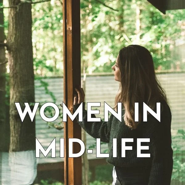 Woman standing in a house, looking out the window toward a forested backyard.