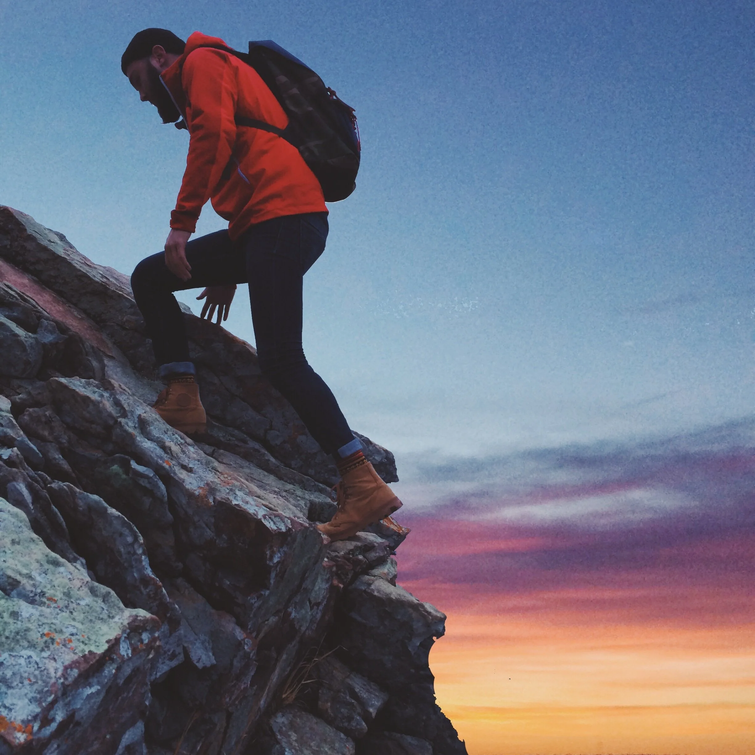 Photo of a man in a red sweatshirt, climbing up a rock at dusk, with a cloudy light blue and pink sky in the background.