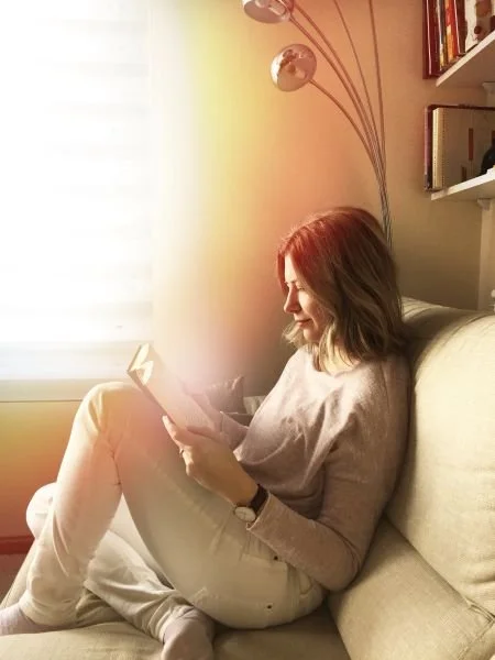 Woman seated on a sofa reading a book in a dimly lit room.