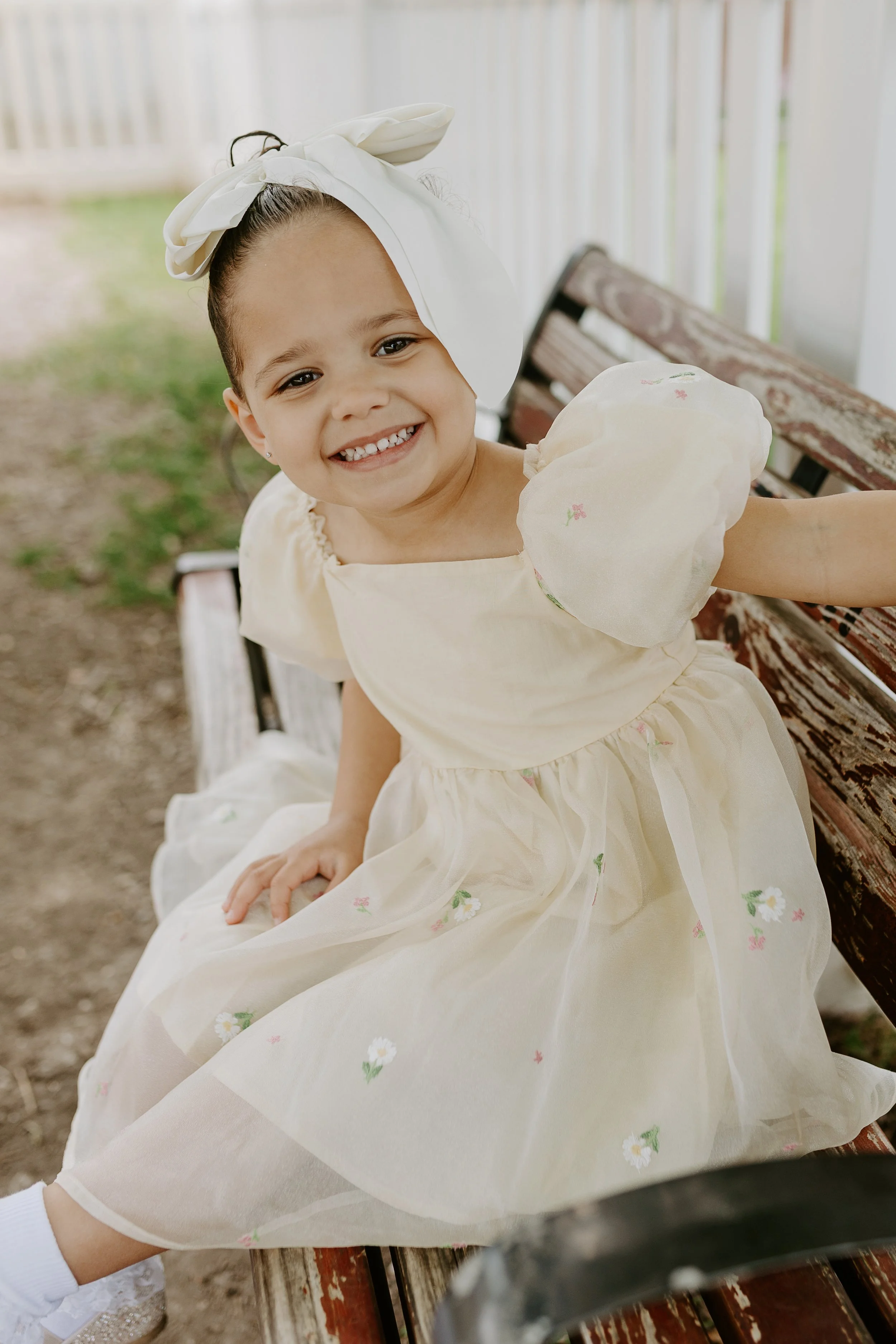 A young girl smiling sitting on a wooden park bench wearing a cream-colored dress with floral embroidery and a large cream bow headband.