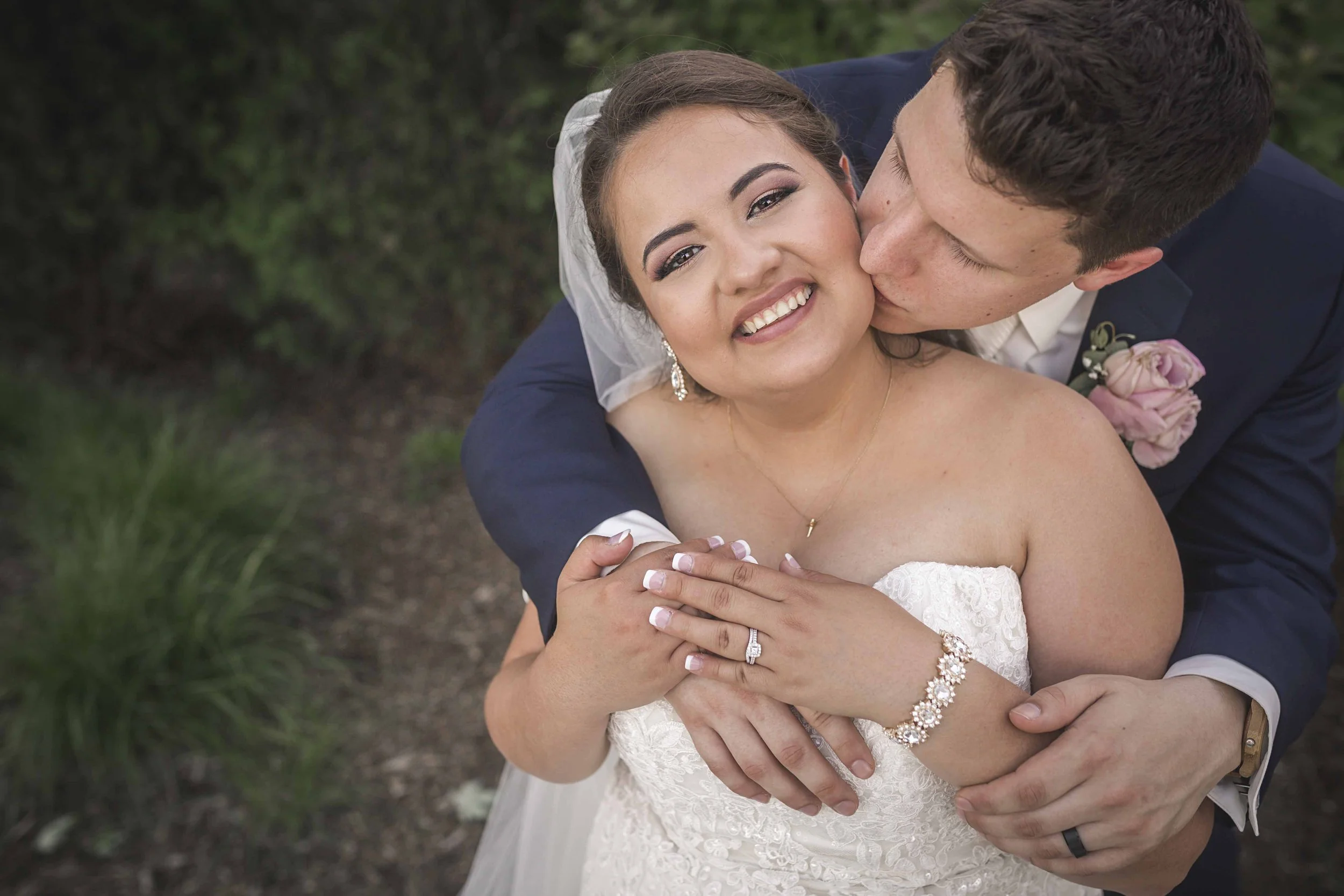 A newlywed couple embracing outdoors, with the groom kissing the bride on her cheek. The bride is smiling, wearing a white lace wedding dress, earrings, a bracelet, and an engagement ring. The groom is in a dark suit with a pink boutonniere. The background includes green foliage.