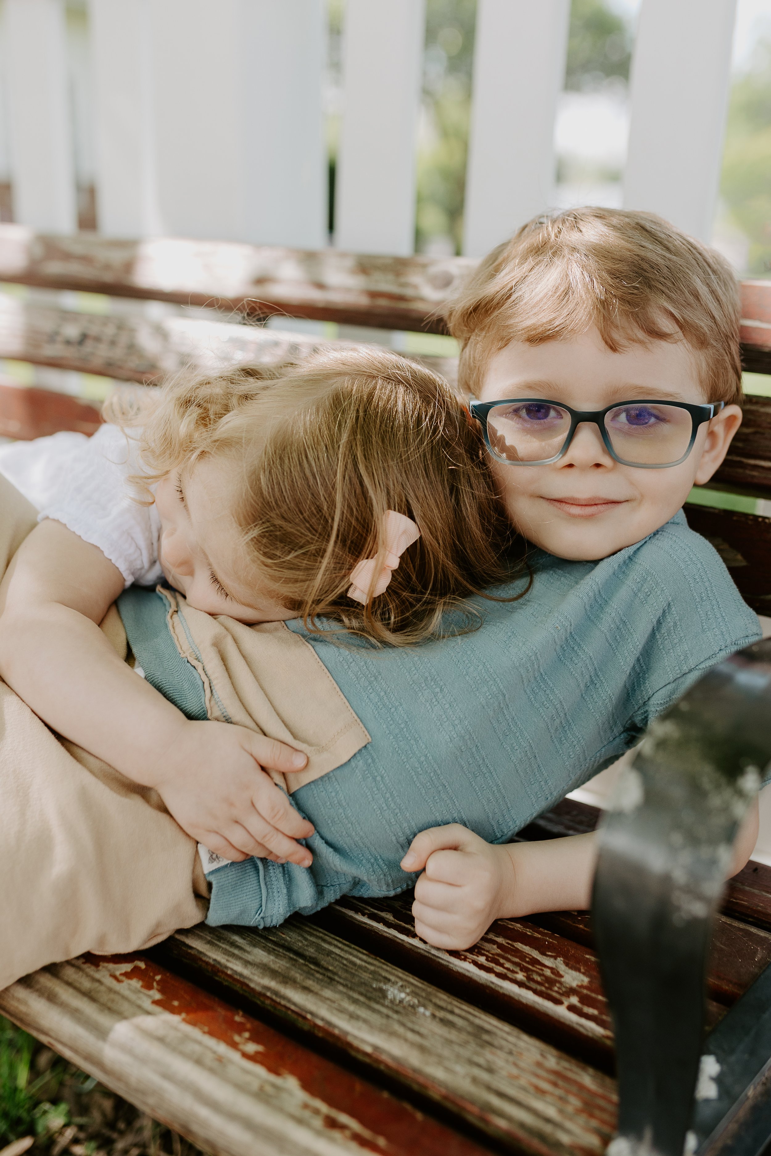 A boy with glasses lying on a wooden park bench with a girl with a pink bow cuddling against his chest.