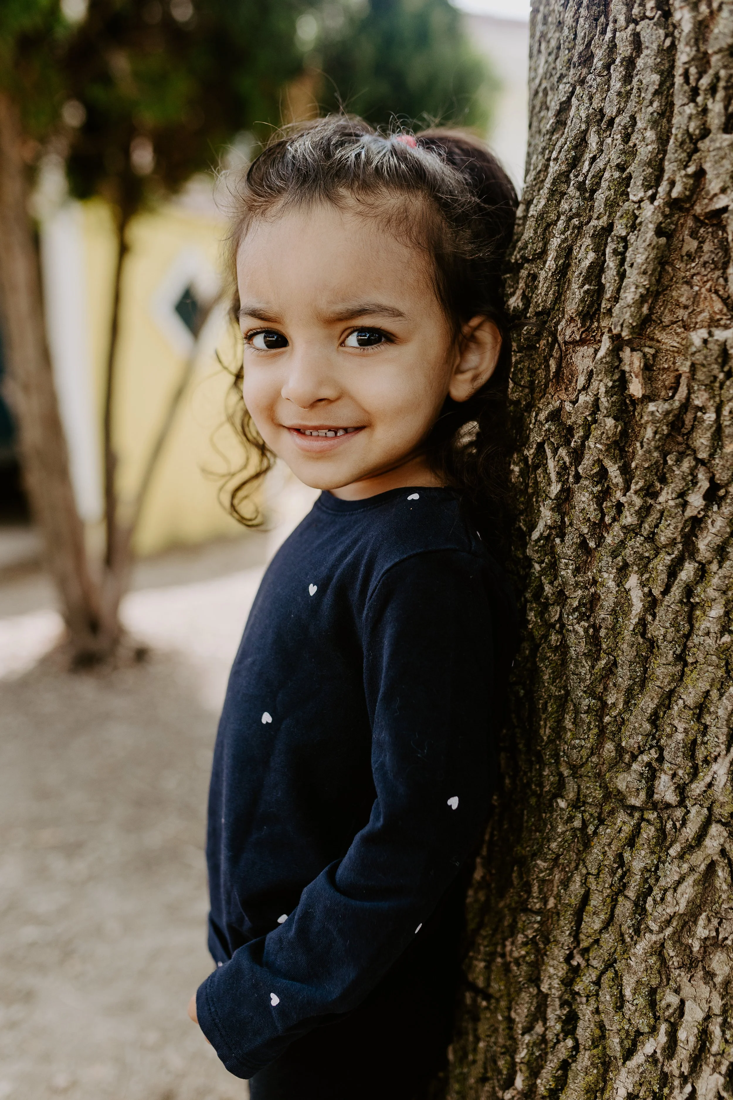 Young girl leaning against a tree outdoors, smiling at the camera.
