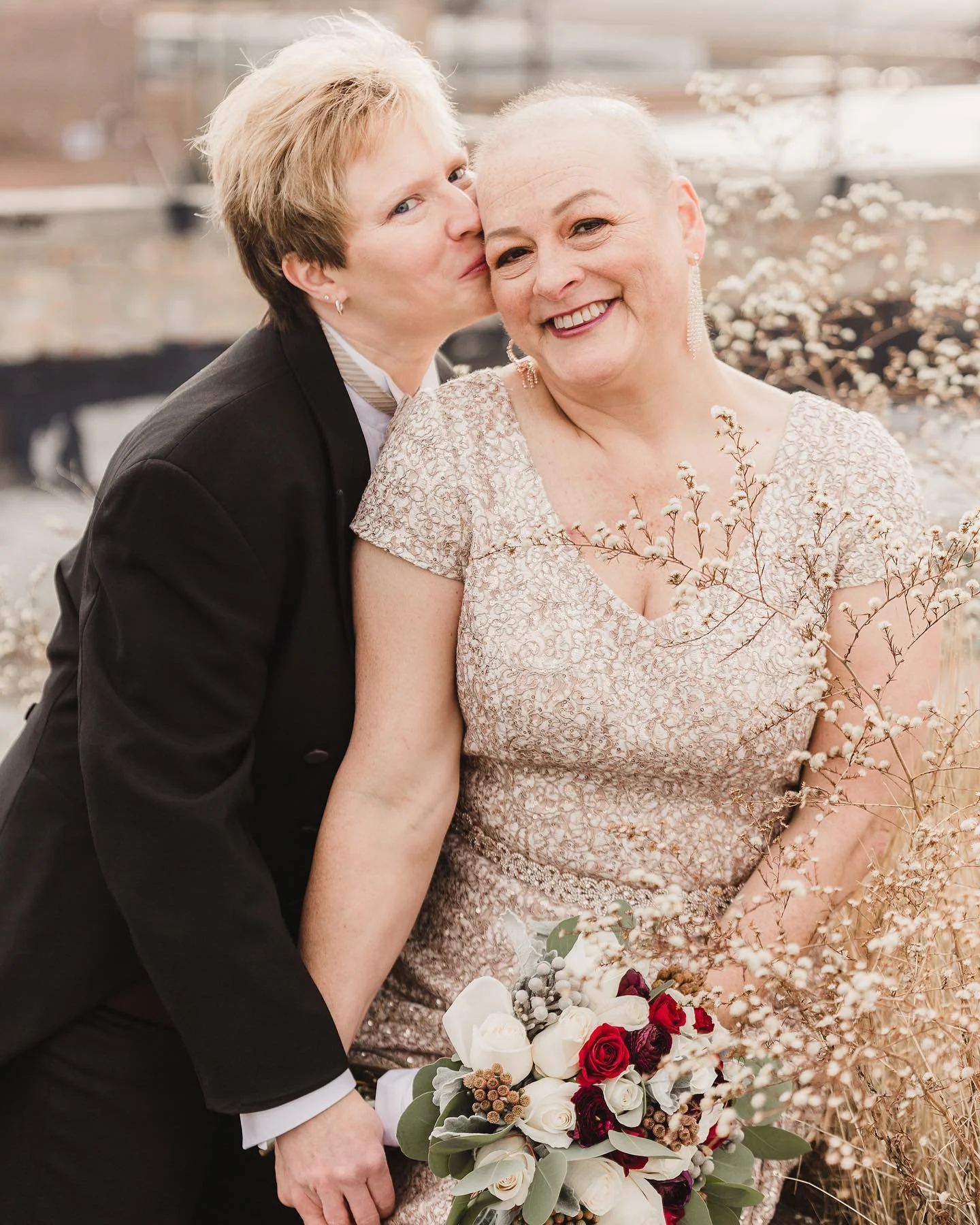 Two women, one with short blonde hair and the other with a shaved head, smiling and embracing each other, surrounded by dried flowers, with one holding a bouquet of roses and other flowers.