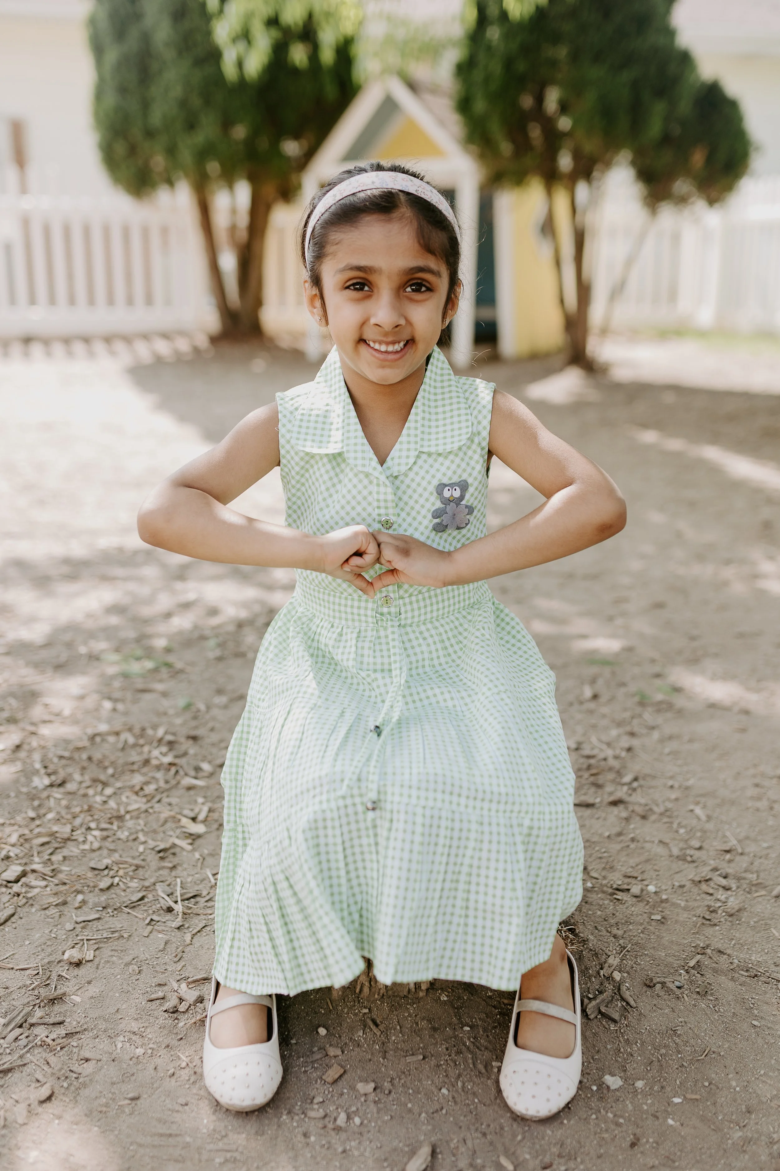 Young girl with dark hair and a headband smiling and making a heart shape with her hands outdoors on dirt ground, wearing a green and white checkered dress with a teddy bear patch.