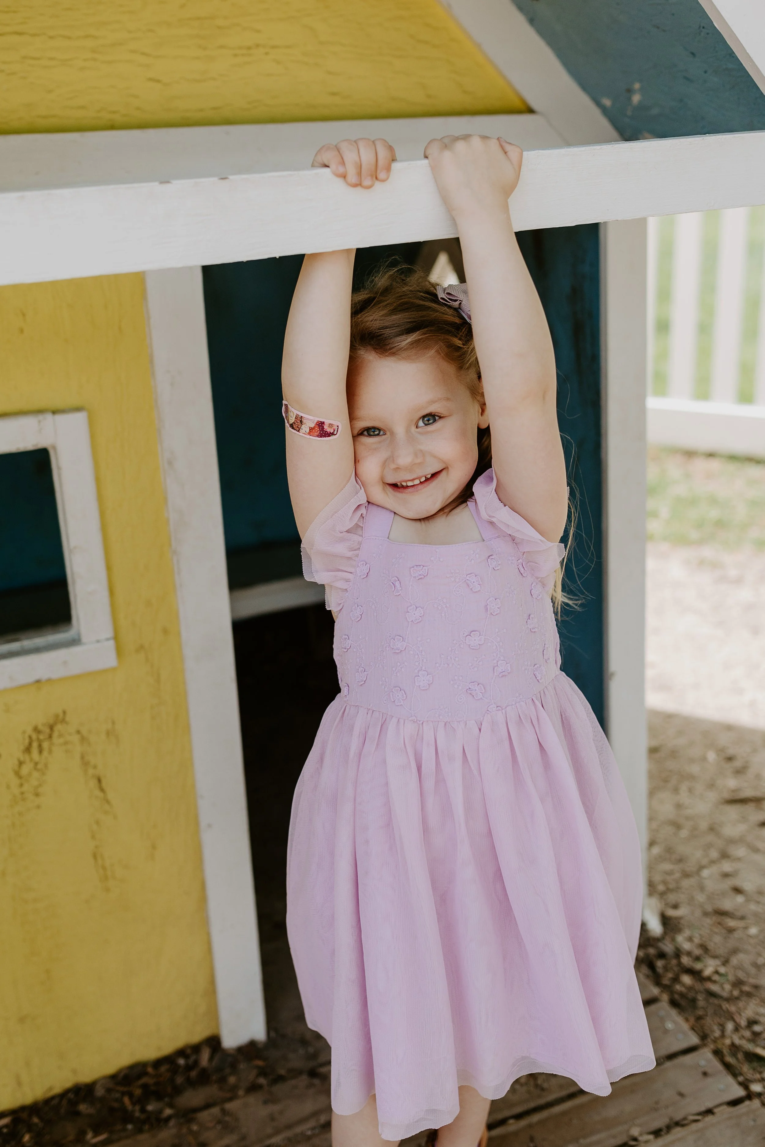 A young girl in a pink dress hanging from a wooden structure outdoors, smiling at the camera.