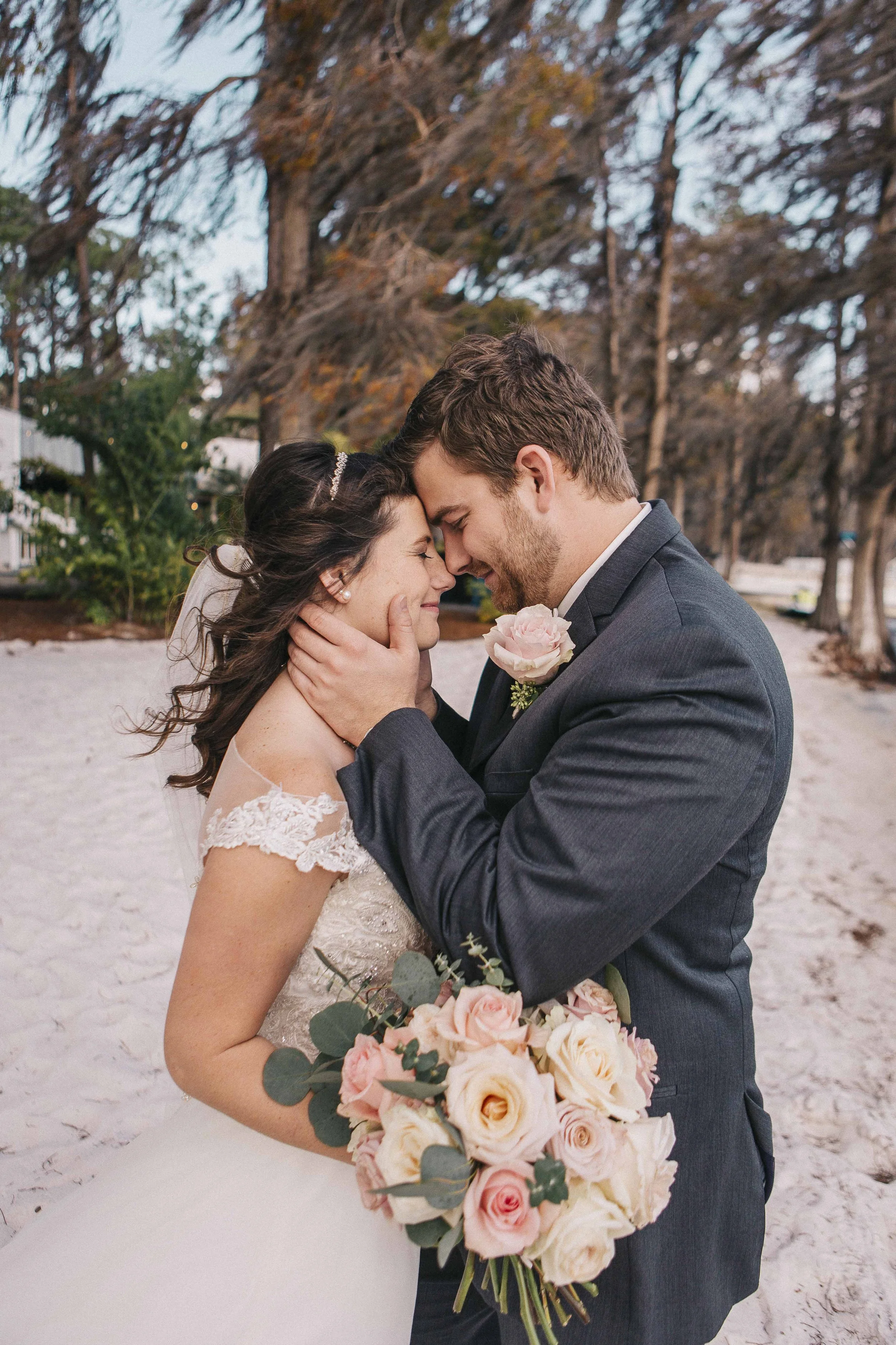 A bride and groom sharing a tender moment on their wedding day, embracing with foreheads touching, outdoors with trees in the background, the bride holding a bouquet of pink and white roses.