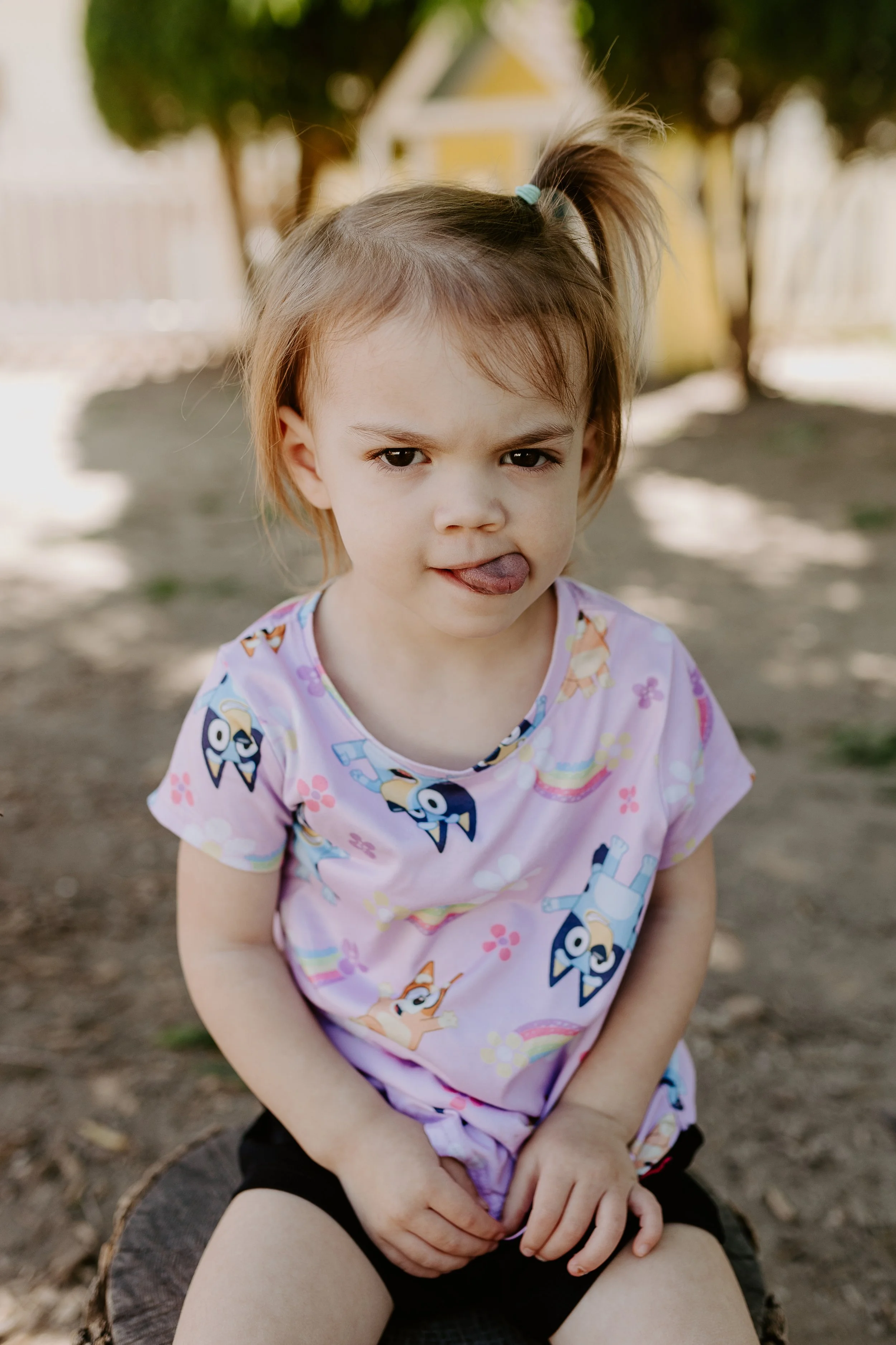 A young girl with light brown hair in a ponytail, wearing a pink shirt with cartoon characters, sitting outdoors on a tree stump, sticking her tongue out.