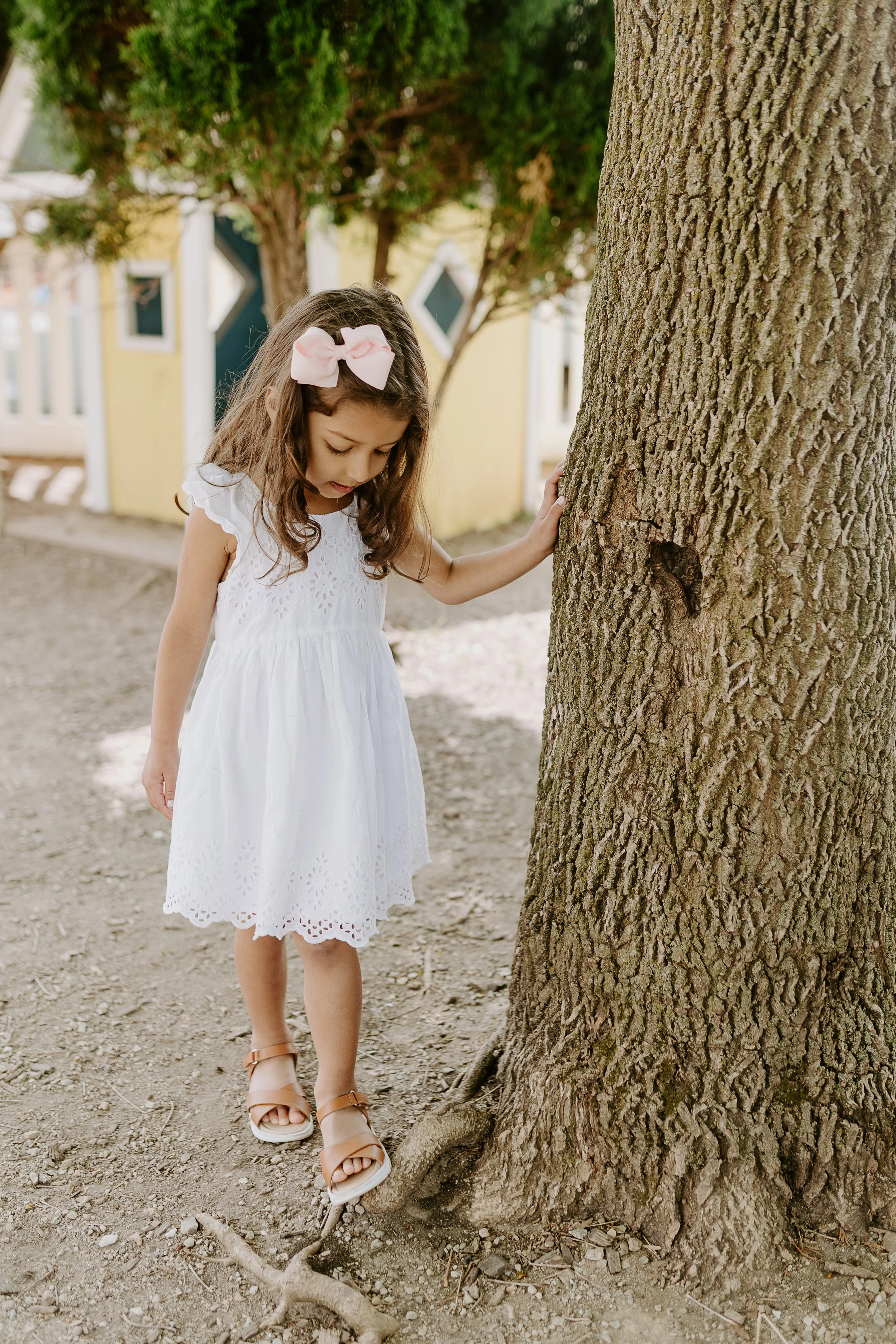 A young girl in a white dress with eyelet embroidery and tan sandals, touching a tree trunk with her right hand, looking down with a curious expression.