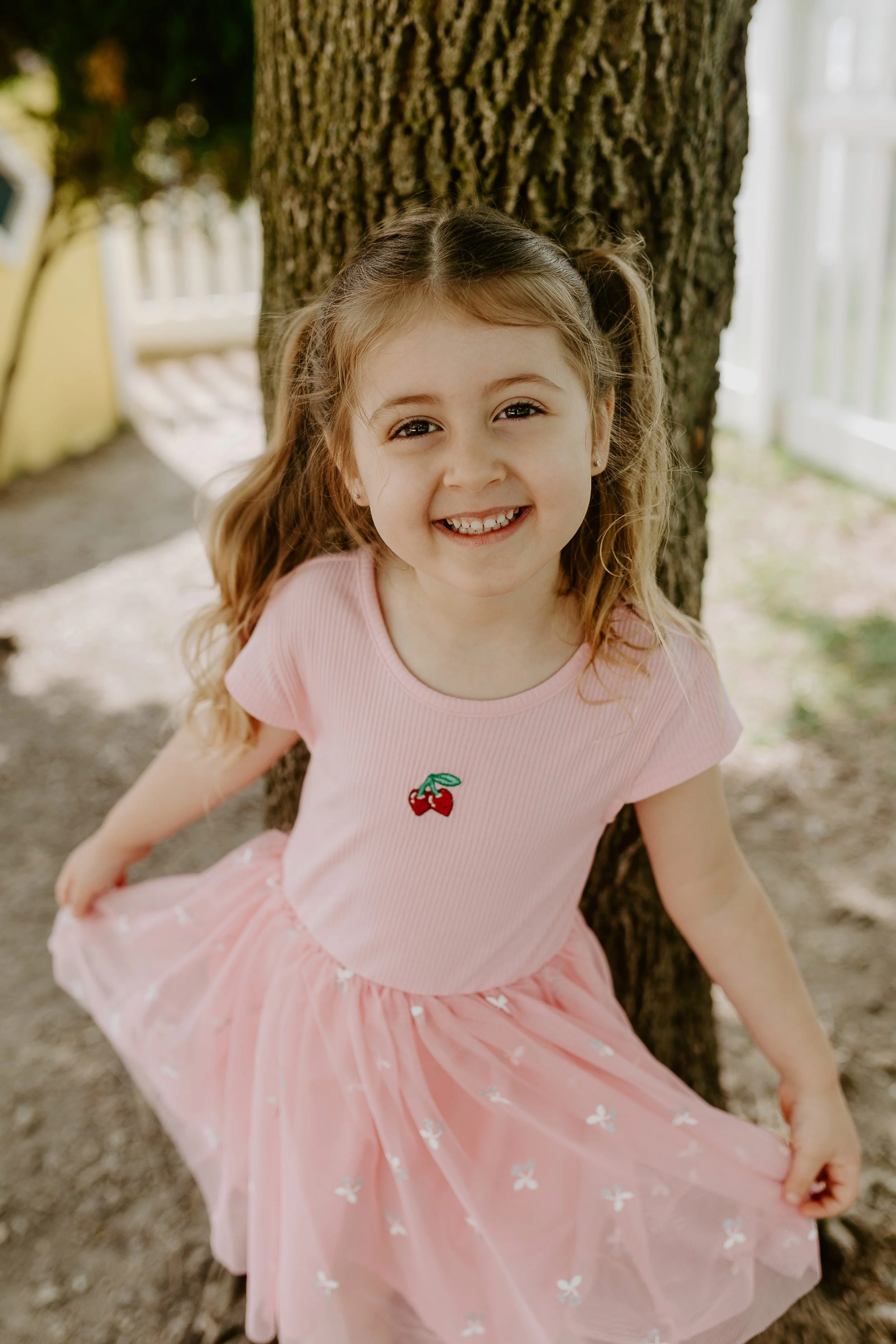 A young girl with long, wavy, light brown hair styled in pigtails, smiling and looking at the camera, standing outdoors beside a tree. She is wearing a pink dress with a cherry embroidery on the chest and a fluffy pink skirt with white flower patterns.