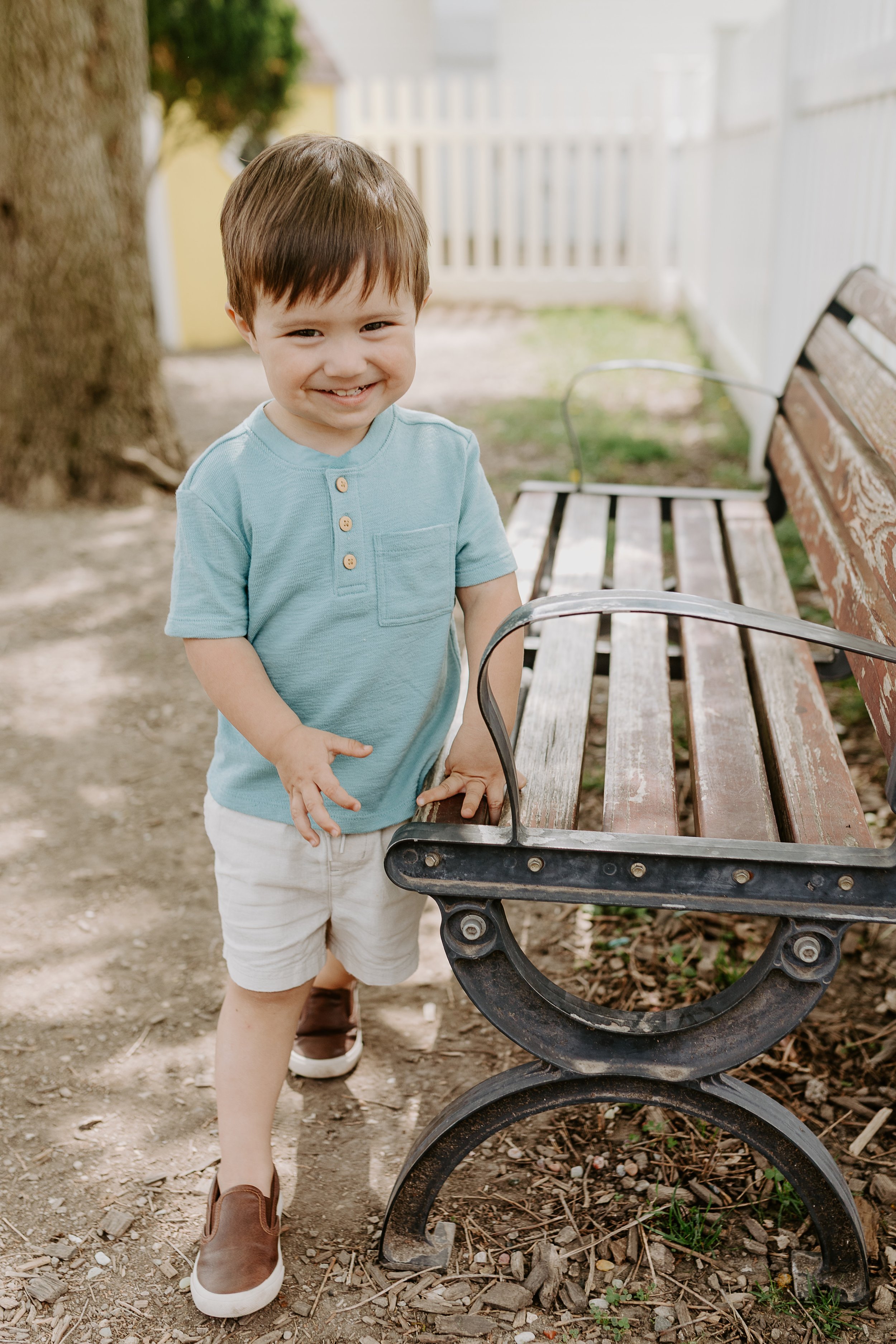 A young boy with brown hair, wearing a light blue shirt, white shorts, and brown shoes, standing outdoors next to a wooden park bench, smiling and holding onto the bench.